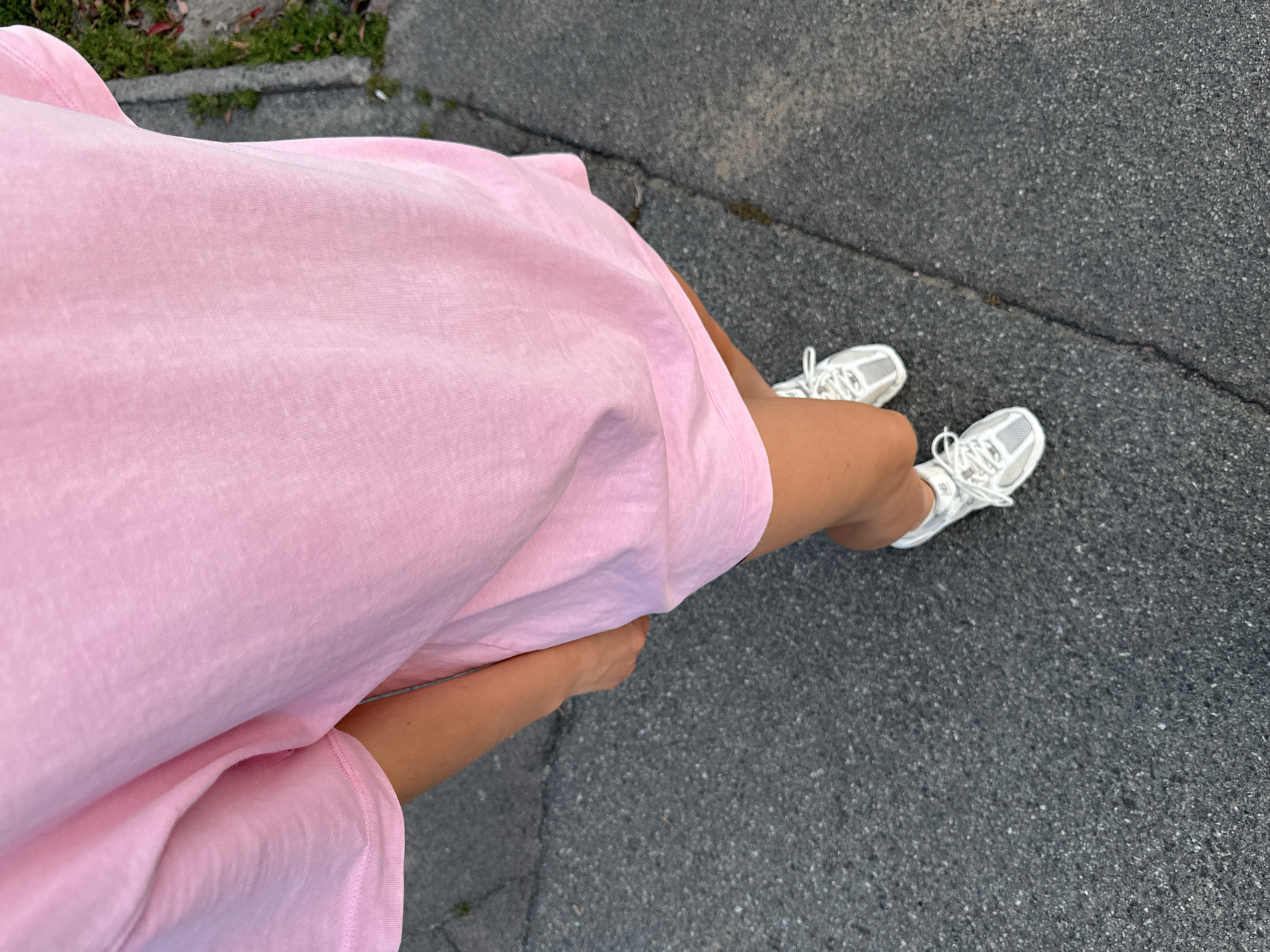 The image shows a close-up, high-angle view of a person standing on an asphalt surface, likely a sidewalk or road. The person is wearing a light pink oversized t-shirt dress and white sneakers. The t-shirt fabric appears to be textured and somewhat creased. One hand is visible, tucked into the side of the dress. The asphalt is dark gray with visible cracks and some scattered small green vegetation along the edge on the right side of the frame. The lighting suggests it is daytime, with shadows potentially indicating late afternoon or early morning. The scene does not contain any specific landmarks or elements that would definitively place it within Kyiv, Ukraine, or any other specific city.