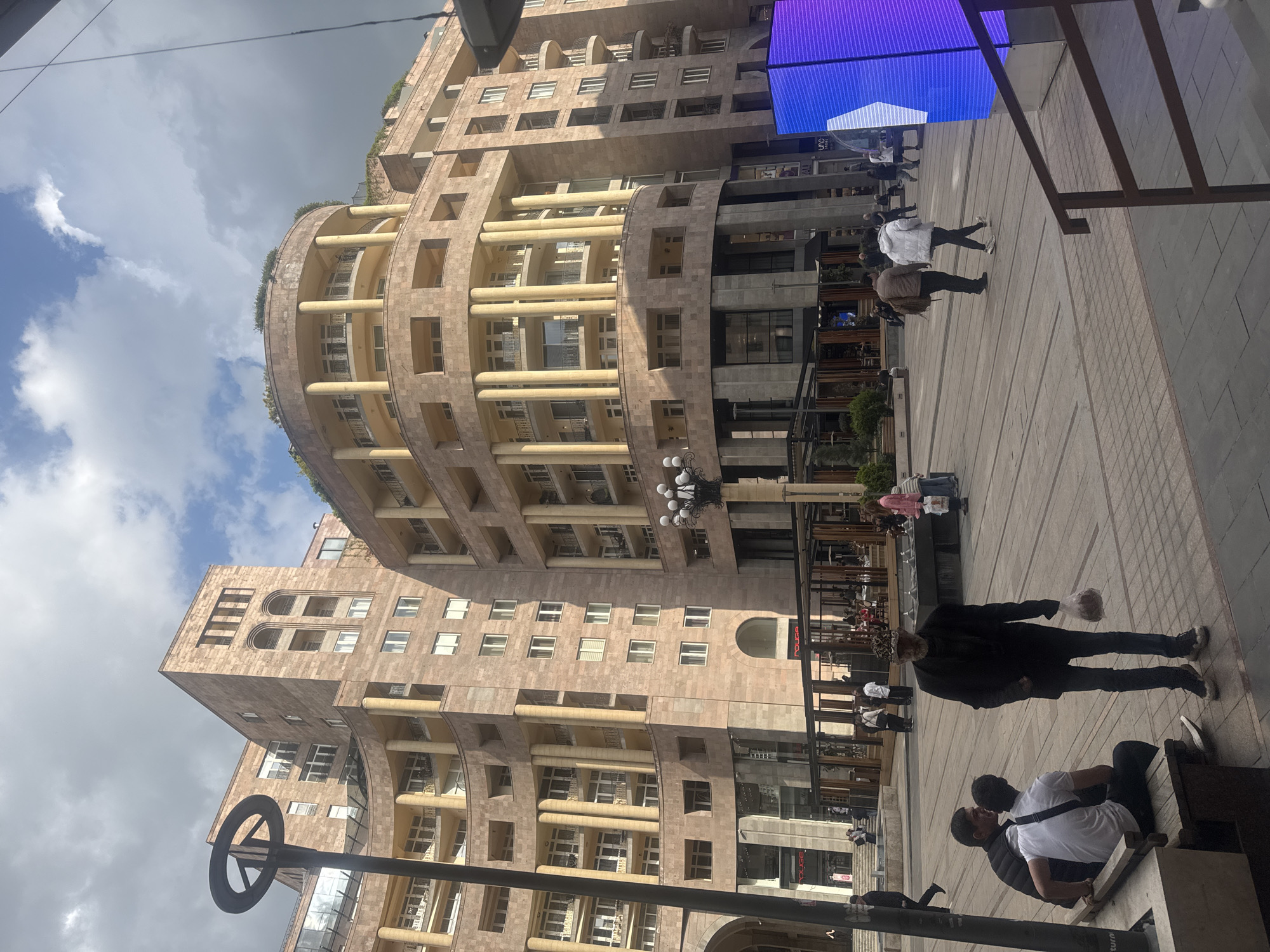 This media file depicts an urban scene in Yerevan, Armenia, showcasing a distinctive multi-story building and a pedestrian area with people.
The prominent subject is a large, modern building constructed from light-colored stone, possibly Armenian tuff. It features a striking design with a curved facade on one section, adorned with numerous balconies supported by columns across multiple floors. Other parts of the building have more angular lines with regularly spaced rectangular windows. Greenery is visible on some of the upper terraces, adding a touch of nature to the architecture.
On the ground level, a wide, paved pedestrian plaza extends, where several individuals are engaged in various activities. In the foreground, two men are seated on a low concrete bench or ledge. Further into the scene, a man in a dark jacket is walking, carrying a plastic bag in his hand. Other people are casually strolling or standing throughout the plaza, suggesting a typical day in a bustling urban environment.
To the right side of the image, a large digital display screen emits vibrant blue and white light, showing abstract graphics. Below the screen are various storefronts. One shop clearly displays the word "POLICE" in red lettering on its sign, likely indicating a brand or store name. Another sign partially visible seems to have "UNO" or similar text.
The scene is captured during the daytime, under a partly cloudy sky with patches of blue. The overall atmosphere is that of a lively city street or commercial square.