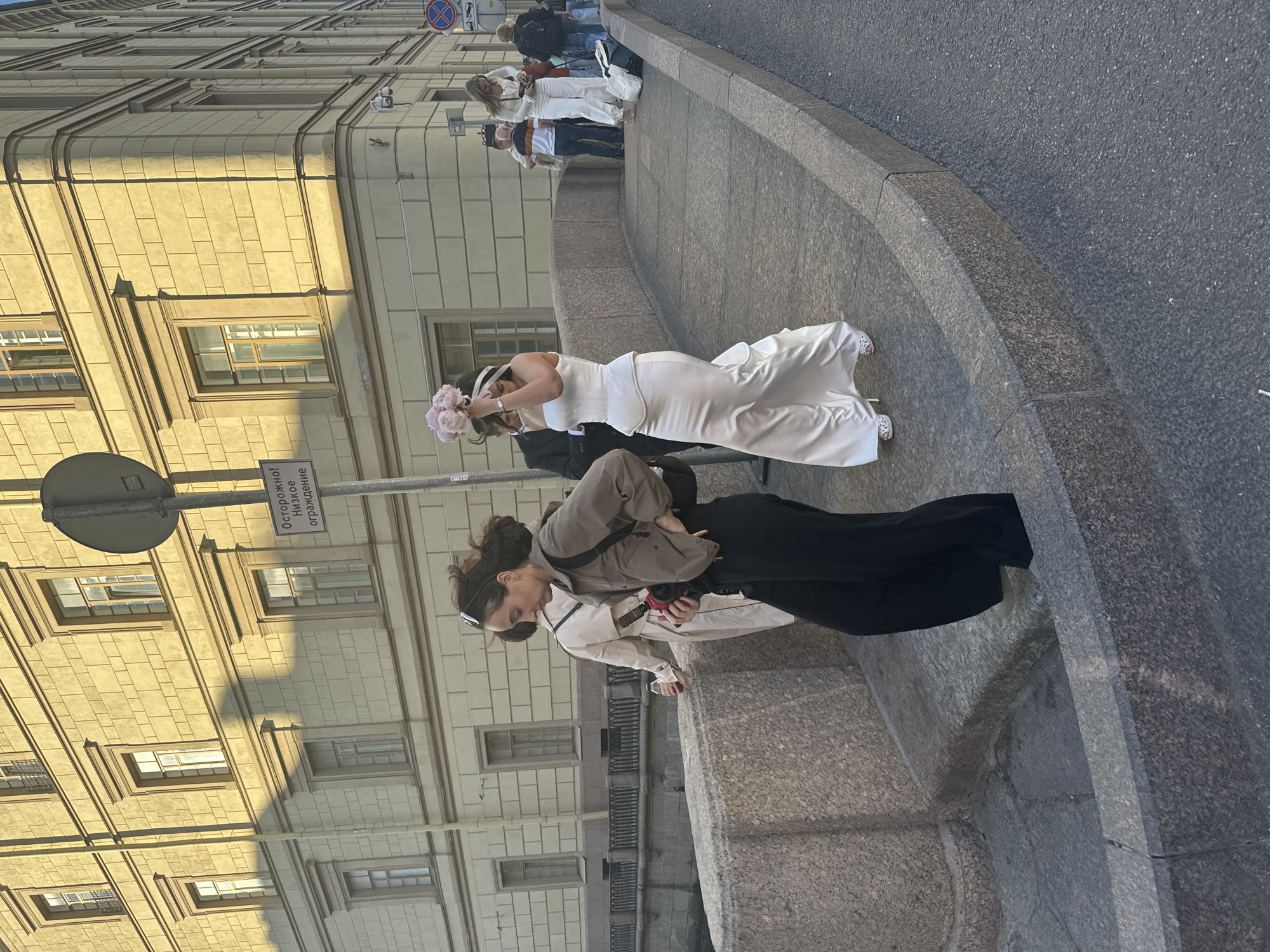 The image shows a street scene in Saint Petersburg, Russia, likely near a canal due to the presence of a stone bridge and railing. The sun is shining, casting long shadows from a building with a neoclassical facade, featuring repetitive windows with decorative surrounds. A sign with Russian text, "ОСТОРОЖНО! НИЗКОЕ ОГРАЖДЕНИЕ," warning of a low railing, is visible on a pole.
In the foreground, a woman wearing a white satin wedding dress with a mermaid silhouette and a bouquet of pink peonies is walking across the bridge. She has dark hair tied back and is wearing white high-heeled sandals. To her left, another woman in a casual olive-green jacket, black pants, and a black backpack is walking in the same direction, holding a camera. Further back, on the right side of the bridge, a woman in white pants and a white top is holding a camera, and behind her, other individuals are visible, including a man in a black suit and another man in casual clothing. The bridge is made of stone, with a textured surface. The street itself is paved with asphalt. The overall scene suggests a post-wedding photo shoot or celebration.