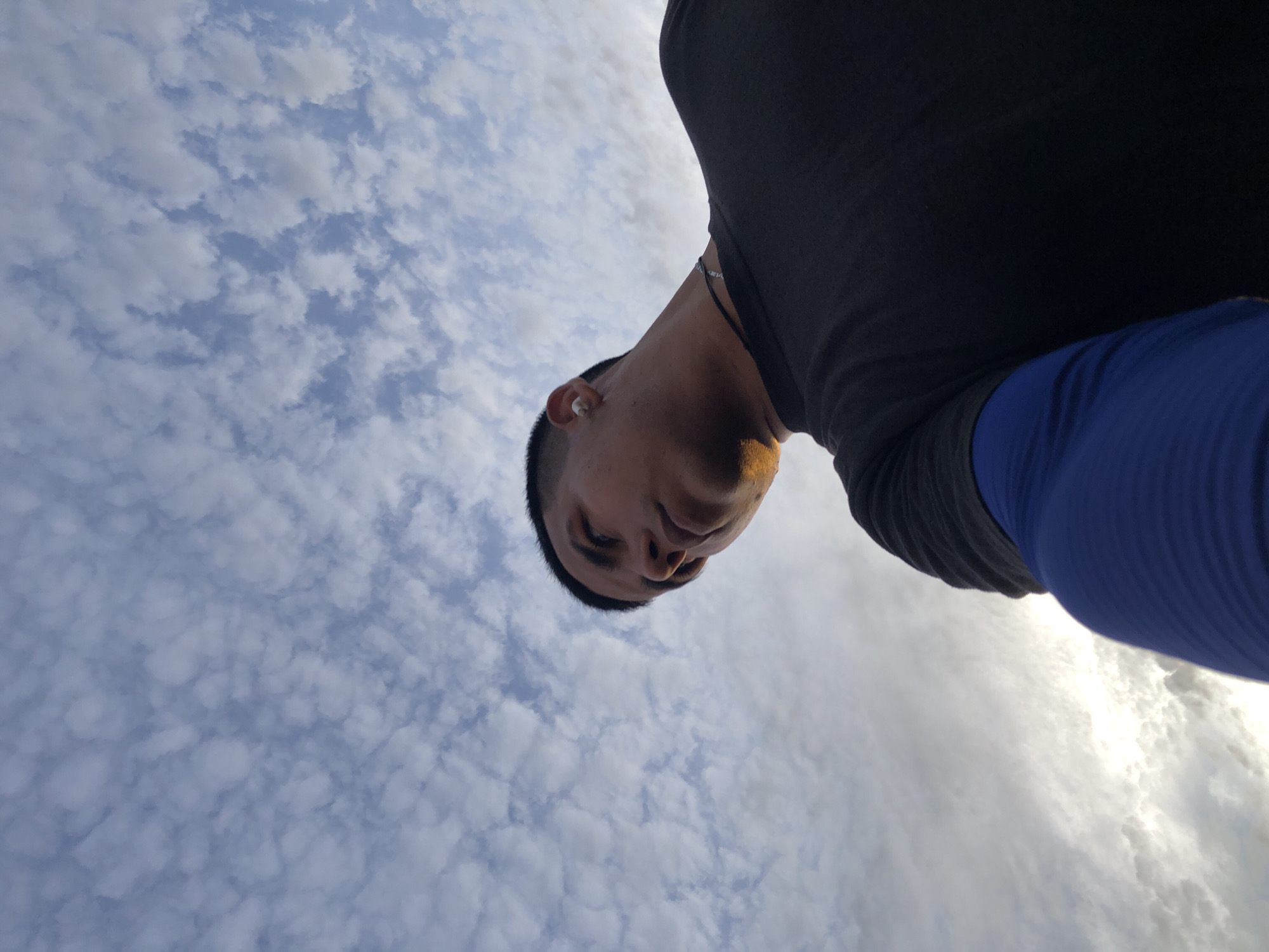 A low-angle shot captures a person looking down at the camera against a backdrop of a sky filled with small, puffy clouds. The person has short, dark hair and appears to be wearing an earbud in their left ear. They are wearing a dark-colored shirt with a blue sleeve visible on the right side of the frame. The sky has a gradient of light blue to white, suggesting sunrise or sunset. The exact location in Juan Griego, Venezuela, cannot be determined without further details or landmarks visible in the image.