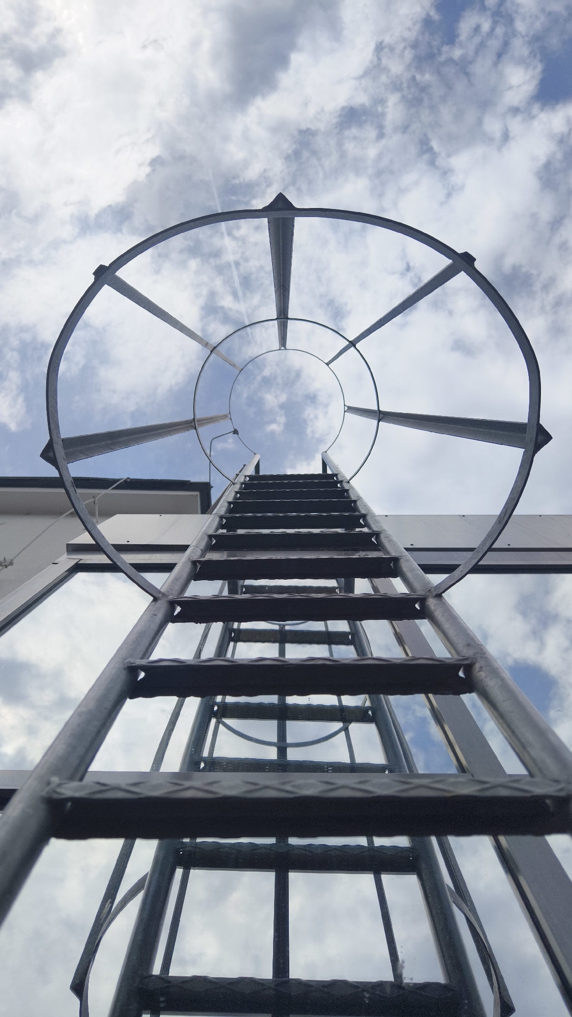 The image shows a low-angle shot looking up at a metal ladder with a safety cage ascending towards a cloudy sky. The ladder is attached to a building with a white facade and large windows that reflect the sky. The scene appears to be on a rooftop or an elevated outdoor area. The metallic structure of the ladder and cage is detailed, with visible rungs and support beams. The sky is filled with a mix of white and grey clouds, with patches of blue visible. There is no indication of people or specific actions taking place. The location is not identifiable as Vienna, Austria.