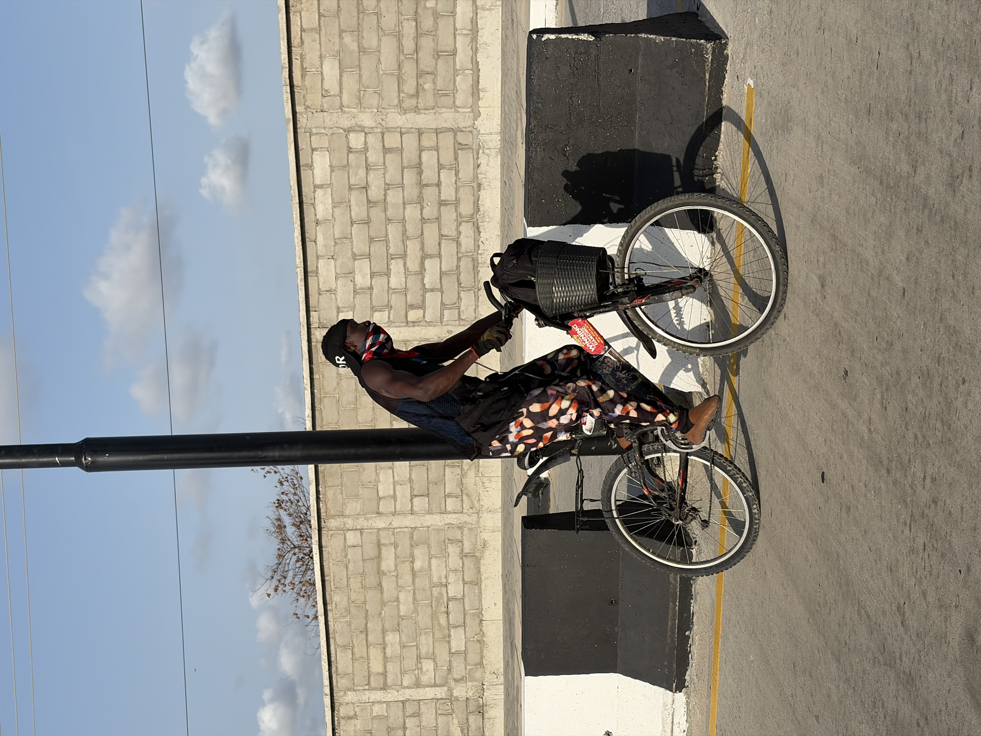 An image shows a man on a bicycle in Igboefon, Nigeria. The man is wearing a black head covering, patterned pants, a blue tank top, and gloves. He is riding a bicycle with a basket on the front. The background includes a brick wall, a black pole, and a partly cloudy sky. The ground is paved with concrete and has a yellow line.