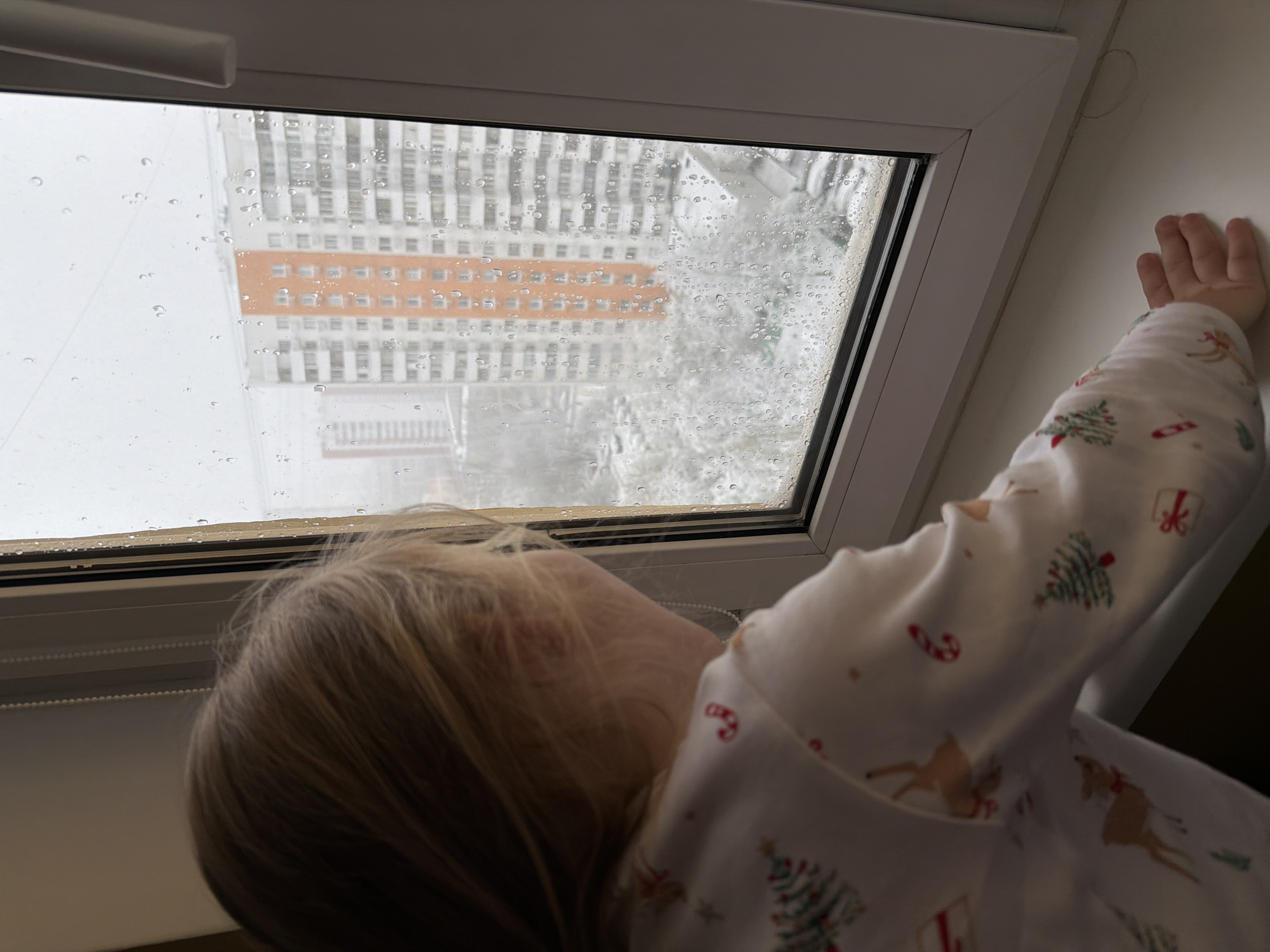 A young child in Christmas-themed pajamas stands near a window in what appears to be an indoor setting. The child's hand is touching the wall next to the window. Raindrops cover the windowpane, partially obscuring the view outside. The visible building through the rain-streaked window suggests an urban environment. Based on the architectural style and available information, the location is likely Khimki, Russia. The building has a distinctive orange section amidst predominantly grey and white facades.