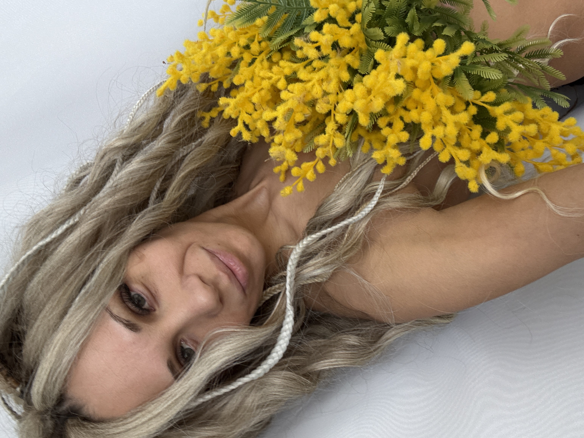 A fair-skinned woman with blonde, wavy hair partially adorned with white braided cords is lying on a white surface. She is holding a bouquet of vibrant yellow mimosa flowers with green foliage. The woman is facing the camera with a slight smile. The location is Unknown, Unknown.