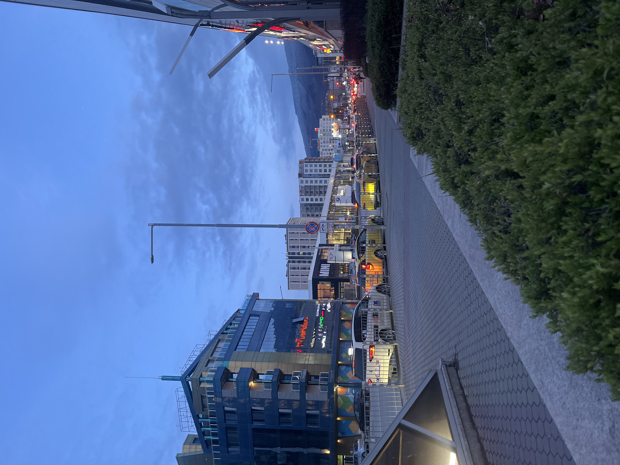The image depicts a street scene in Sofia, Bulgaria, at dusk. Tall buildings line the street, one with a prominent illuminated sign. Traffic is visible with multiple car lights and traffic lights. A mountain range is visible in the distance, under a cloudy sky. A paved sidewalk runs along the right side of the image, bordered by a dense green hedge. Streetlights illuminate the scene. The location appears to be near Vitosha Boulevard, based on the architecture and mountain view.