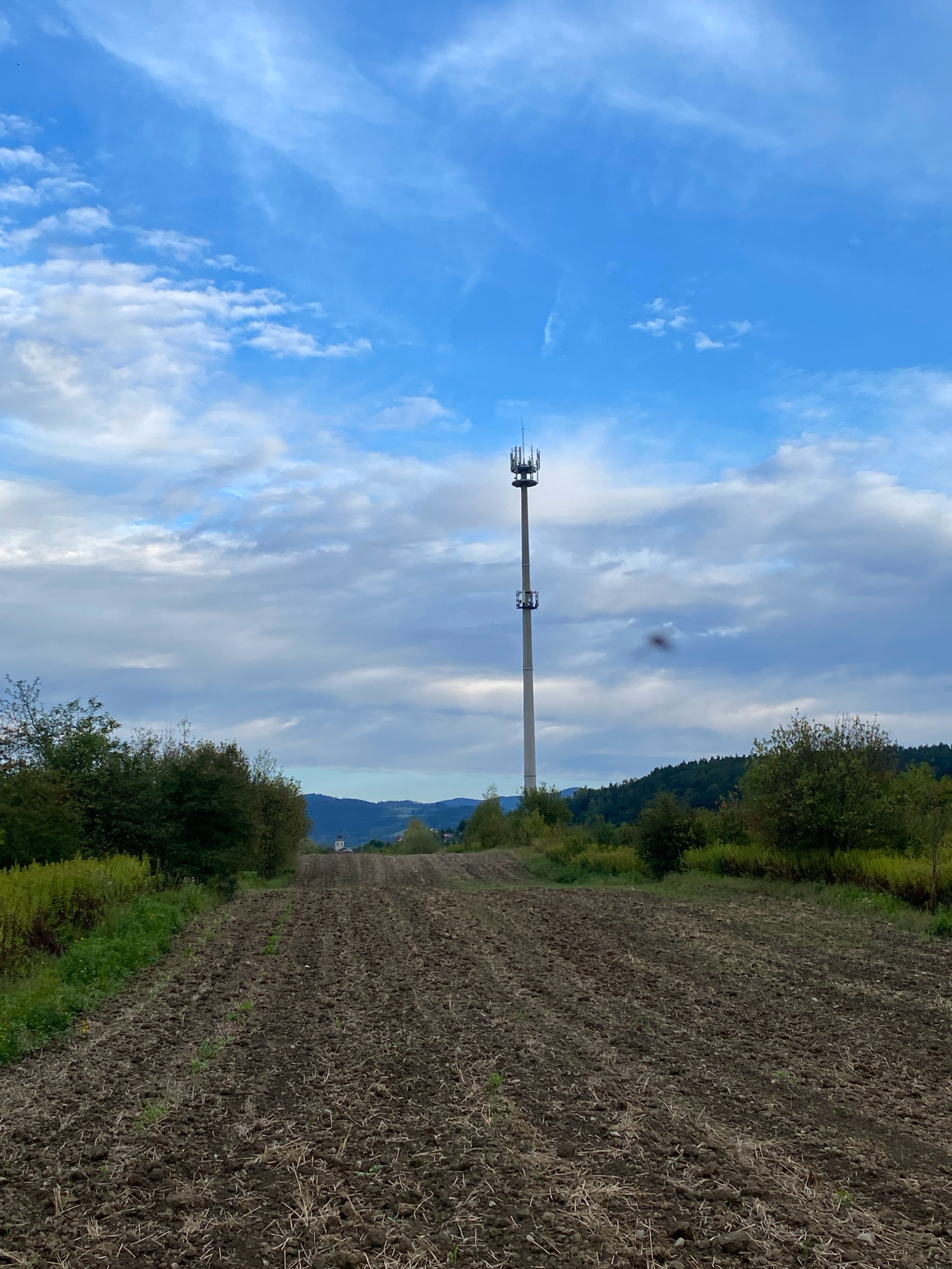 The image depicts a rural landscape under a partly cloudy sky. In the foreground, a plowed field with dark soil stretches towards the horizon. The field is bordered by green foliage and trees on either side. A tall, slender telecommunications tower stands prominently in the middle ground, equipped with antennas at its top. Beyond the field and trees, rolling hills covered in dense green forest recede into the distance, with a small village or settlement visible on the far hillside, including what appears to be a church steeple. The sky is a mix of bright blue and scattered white and gray clouds. The overall scene is one of open countryside with visible infrastructure.