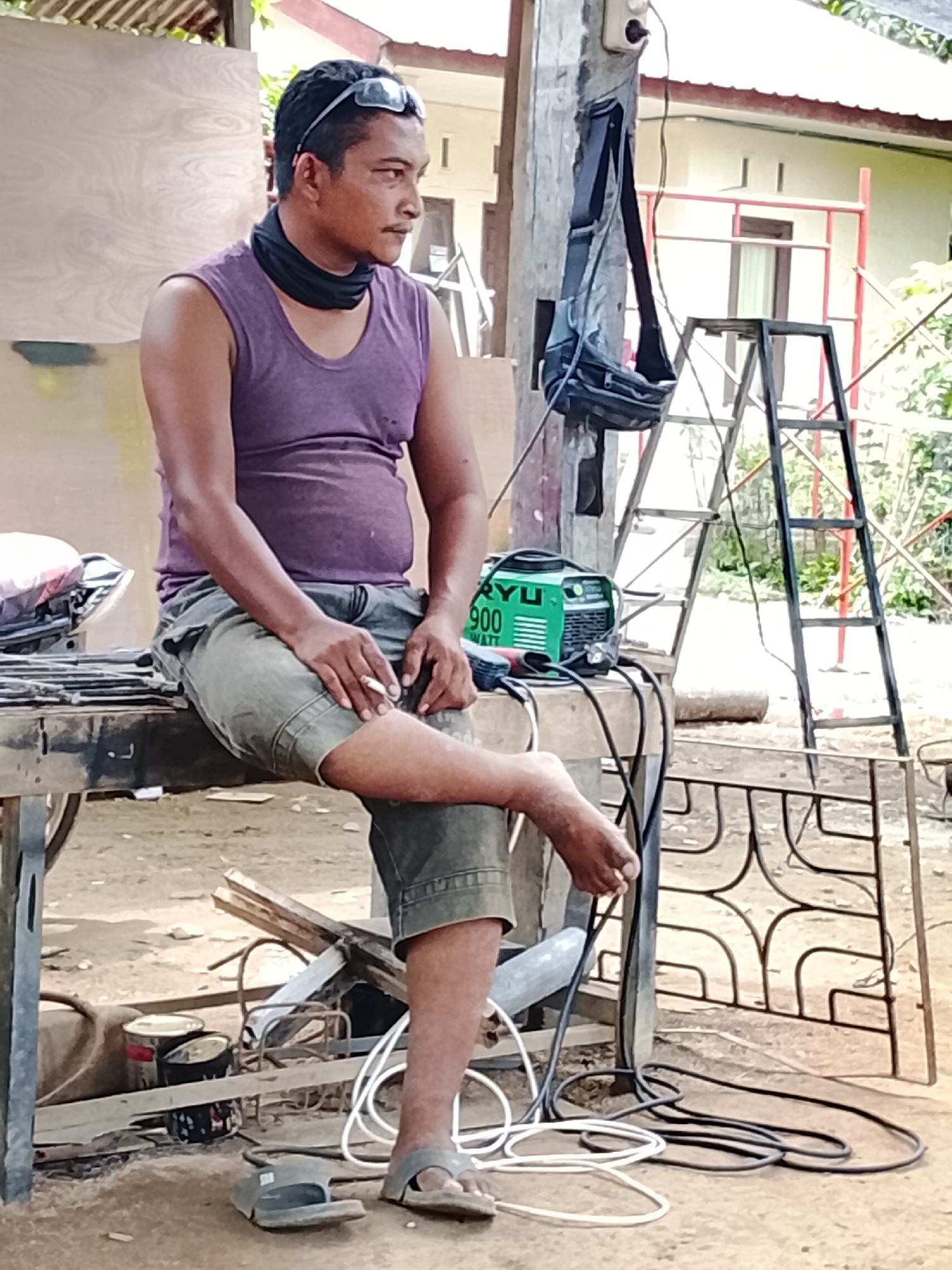 In Mee, Indonesia, a man sits on a wooden workbench. He is wearing a purple tank top, green shorts, and sandals. He has sunglasses on his head and a black bandana around his neck. He is holding a cigarette in his right hand and appears to be smoking. A green welding machine with the label "RYU 900 WATT" is on the workbench to his right. The background shows a ladder, some scaffolding, and a building with a red roof. The ground is dirt and there are several objects scattered around, including cans and welding equipment.