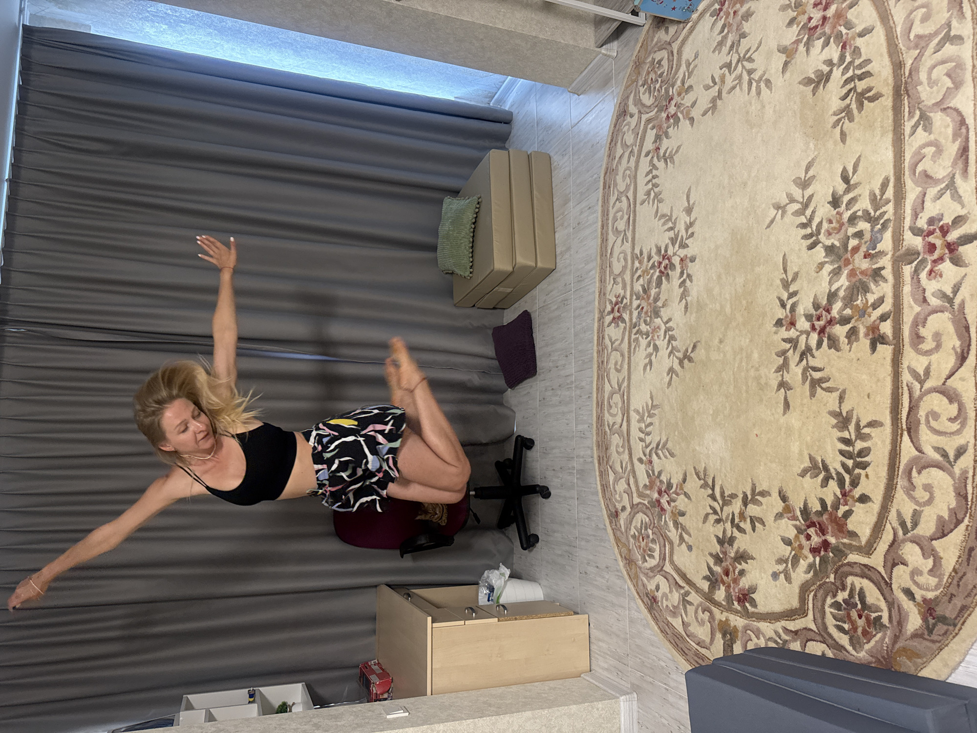 An adult female with blonde hair is captured mid-air, jumping in a room. She is wearing a black crop top and patterned shorts. Her arms are outstretched, and her legs are bent. The background features gray curtains, a desk with a chair, and a large oval rug with a floral pattern. There are also some folded cushions on the floor. A cat is visible sitting on the chair beneath the jumping woman. The flooring appears to be light-colored laminate or wood.