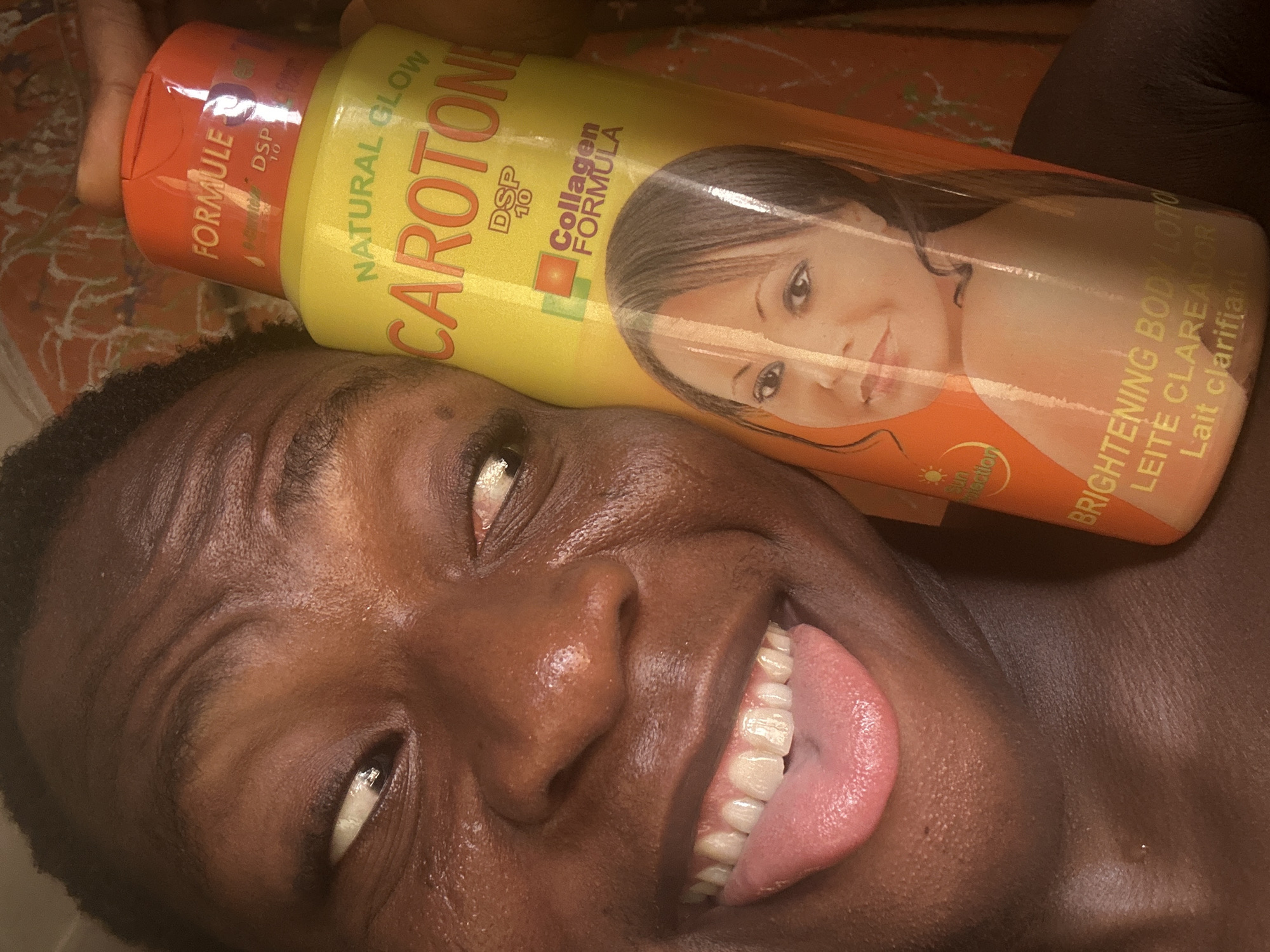 A man with dark skin is posing for a close-up shot while holding a bottle of Carotone Brightening Body Lotion on his head. He is sticking his tongue out and smiling. The lotion is yellow and orange with a picture of a woman on the label. The location is Unknown, Unknown.