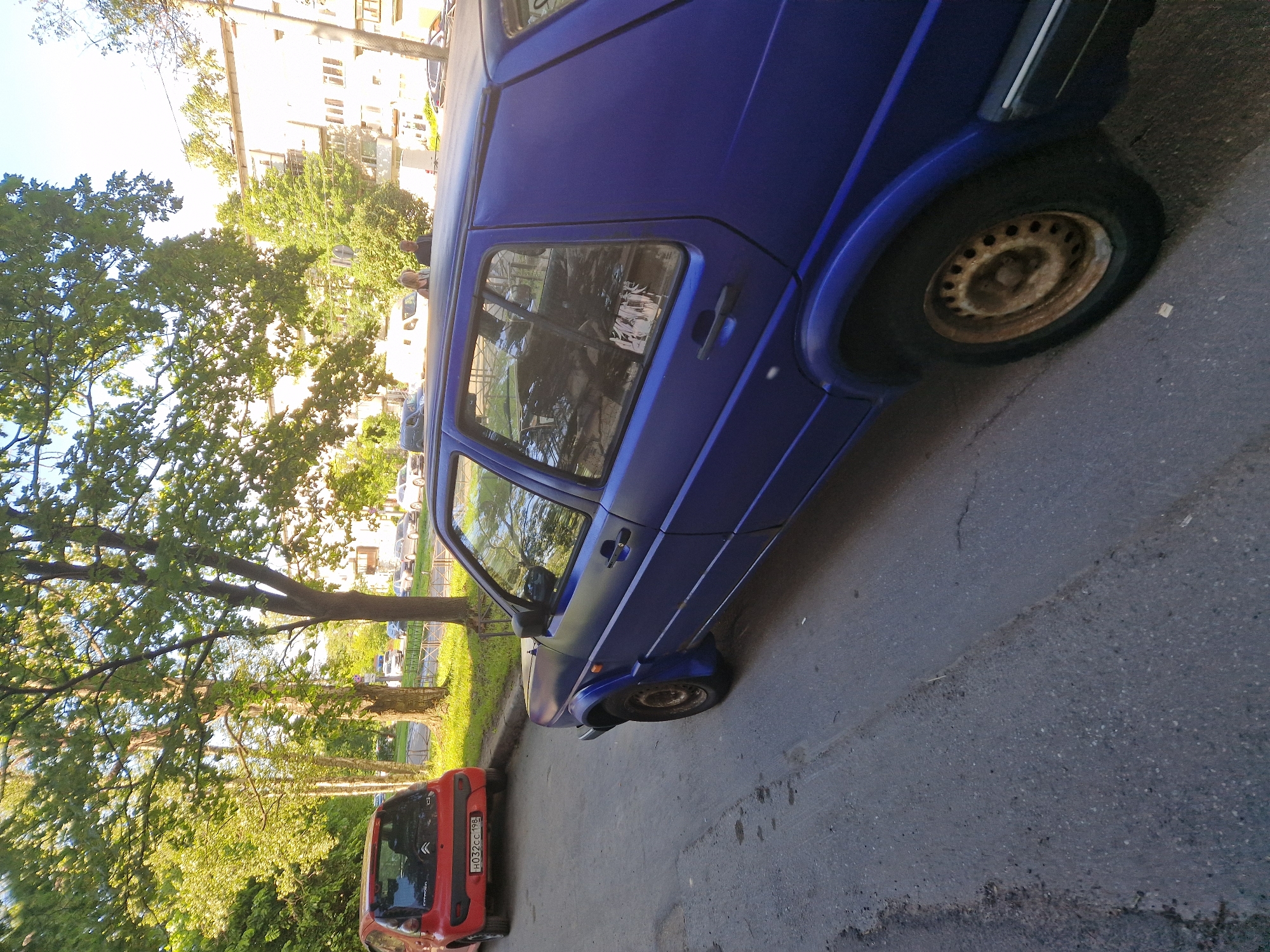 In a city environment, a dark blue, matte-finished, four-door Volkswagen Golf is parked on the right side of an asphalt road. The car is positioned at an angle, with its rear facing away from the camera and slightly towards the left. The driver's side of the car is visible, showing the rear door, window, and fender. The car has a black roof. In the background to the left, a small, red hatchback is parked on the side of the road. The red car's license plate is visible. Behind the cars, a street is lined with trees and a multi-story building is visible further in the background. The sky is clear and bright.