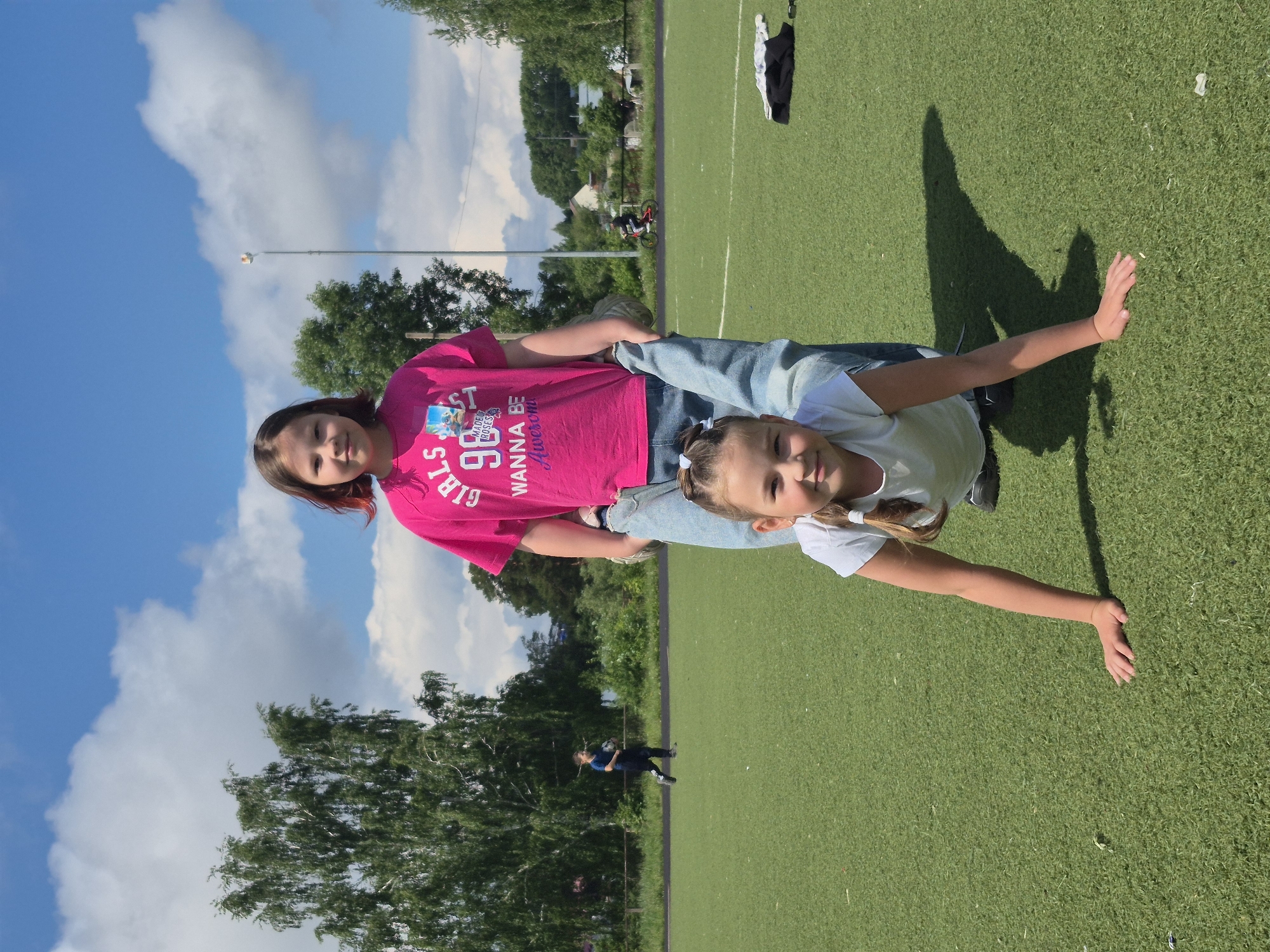 This image, when correctly oriented, captures two girls standing on a green artificial turf field under a bright blue sky dotted with white clouds in Chaikovsky, Russia. The clear daylight is evident from the well-lit scene and the distinct shadows cast by the girls on the field, suggesting it's mid-day or early afternoon.
The older girl, with dark hair, is positioned behind the younger girl. She wears a fuchsia pink t-shirt that displays text including "GIRLS", the number "98", "WANNA BE Awesome", and "MADE OF ROSES", paired with light-colored jeans. The younger girl, with light brown hair tied in pigtails, stands in front of her, dressed in a white t-shirt and light-colored jeans, with her arms outstretched to her sides as if balancing or posing. Both girls are looking towards the camera with slight smiles, conveying a happy and relaxed mood.
The setting is an outdoor sports field, possibly a school or community recreation area. In the background, beyond the field, there are lush green trees and some distant buildings. A person on a bicycle can be seen further in the background, moving along what appears to be a track or path. On the field itself, near the edge, a black and white item (likely clothing) and a small white object are discernible.