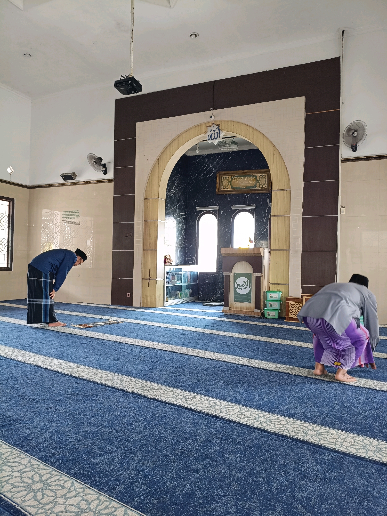 The image depicts the interior of a mosque in Maguwoharjo, Indonesia. Two men are present, both engaged in prayer. The man on the left, wearing a blue garment and a black skullcap, is in a bowing position. The man on the right, in a grey top and purple sarong, is in a similar bowing posture. The mosque features a blue carpet with white and grey striped prayer mats. An arched entryway with dark blue marble walls is visible in the background, leading to a room with windows. Decorative Islamic calligraphy adorns the wall above the arch. A projector hangs from the ceiling, and fans are mounted on the walls. The environment is clean and well-lit, with the sunlight streaming in through the windows.