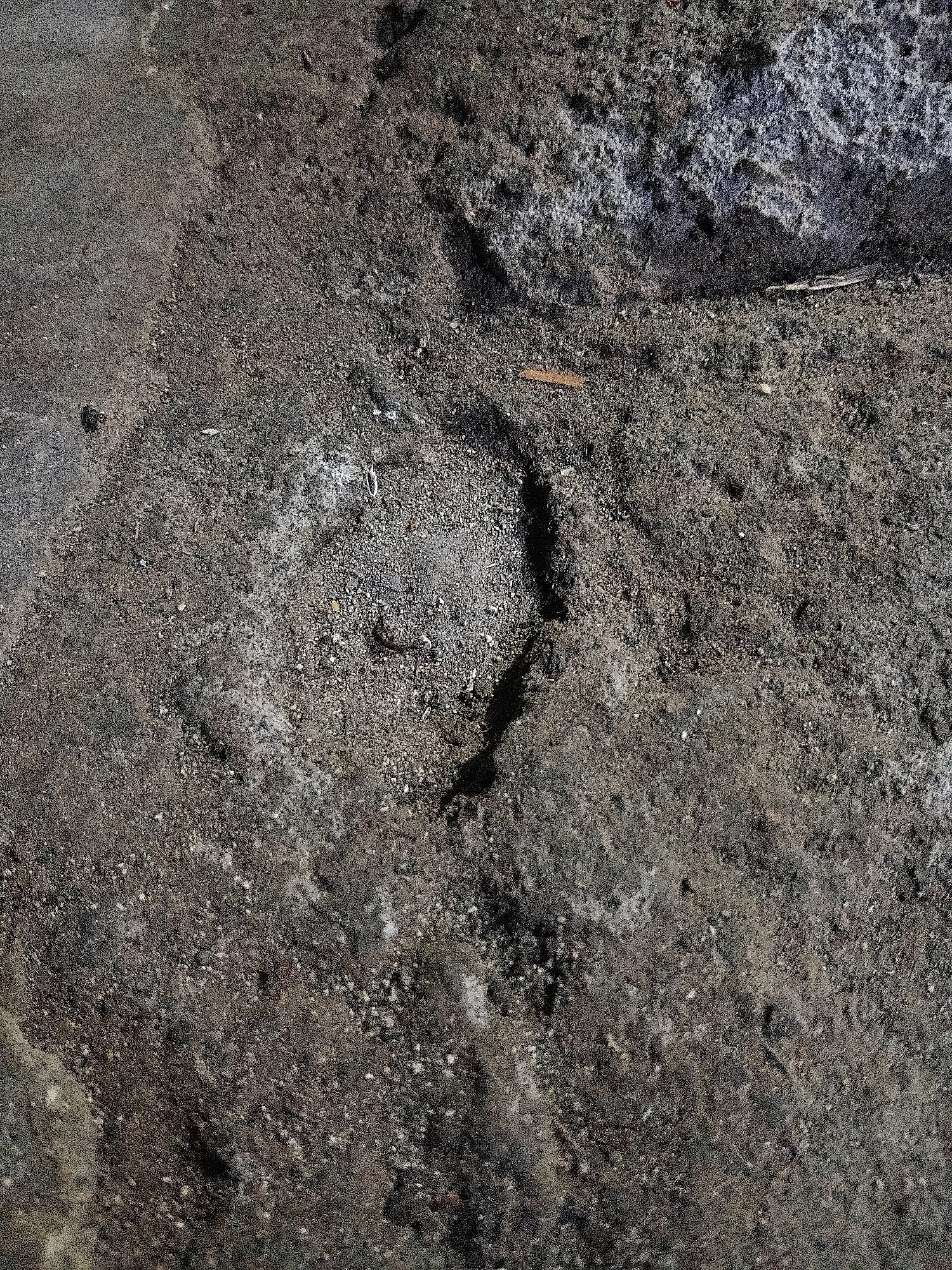 The image displays a close-up view of cracked, dark, granular soil. The texture is rough and uneven, with small pebbles and dust visible. A thin, dark line, possibly a root or a crack in the soil, runs vertically through the center of the image. There are some lighter-colored patches, perhaps mineral deposits or dried residue, scattered across the surface. In the upper right quadrant, there is a lighter, rougher textured area that appears to be a different type of material, possibly stone or compacted earth. The lighting suggests an outdoor environment, with shadows indicating the direction of illumination. The scene is devoid of any human presence or recognizable landmarks that would indicate a specific location within Cikal, Indonesia.