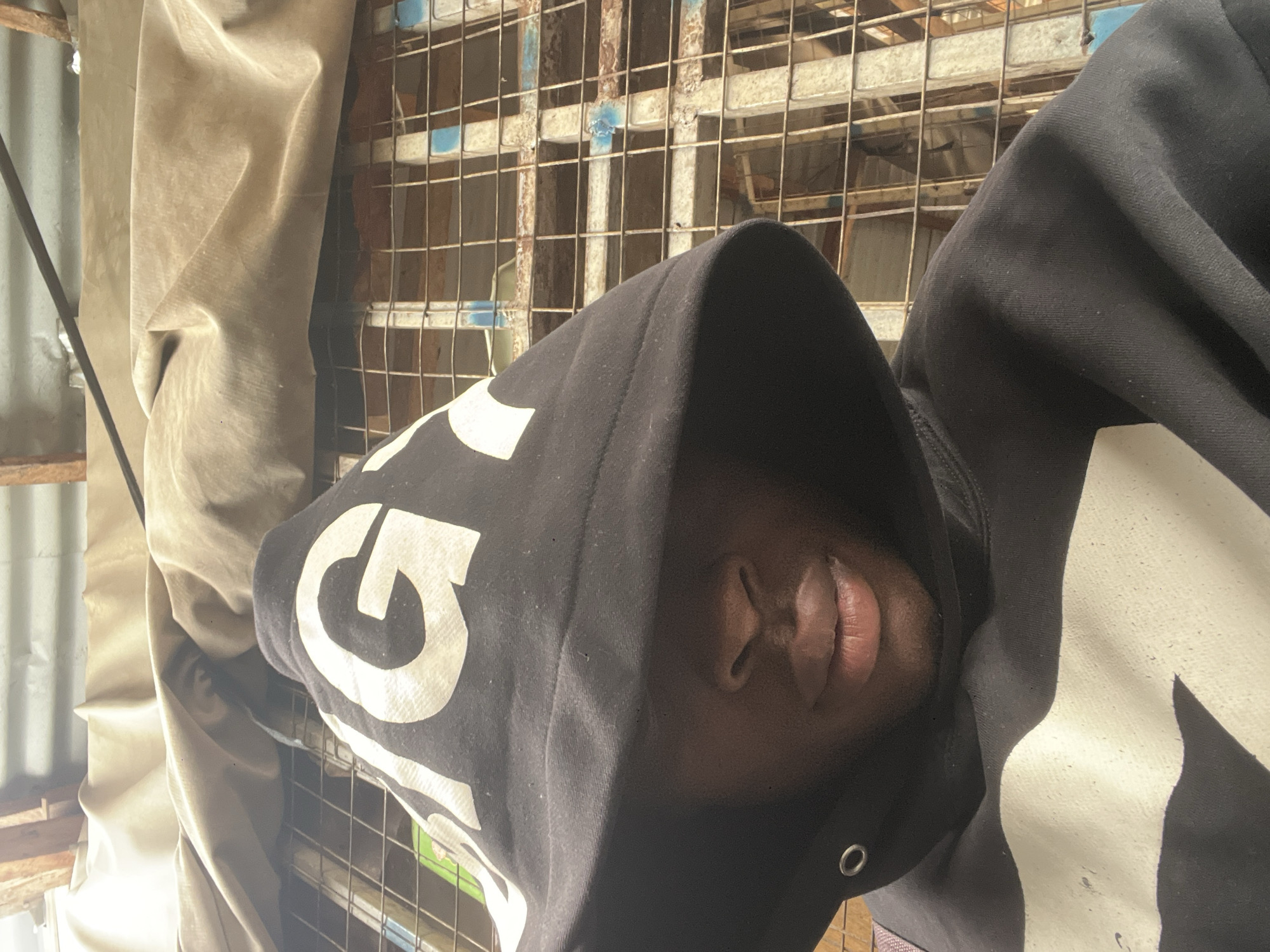 A close-up, rotated portrait photograph depicts a person, likely male, wearing a black hooded sweatshirt. The image is oriented 90 degrees clockwise from an upright view. The subject's head occupies the left side of the frame, with only the lower half of their face – nose, full lips, and chin – clearly visible. Their skin tone is dark.
The black hoodie features prominent, stylized white graphics. A partial letter, resembling a "G," is visible on the upper left side of the hood, while another large, abstract white shape adorns the lower right portion of the garment, possibly on the shoulder or arm.
The background suggests an informal, possibly semi-sheltered outdoor setting in Jalingo, Nigeria. To the immediate right of the person, a weathered metal grid or fence structure is prominent, showing signs of rust and traces of light blue paint. Beyond this grid, other indistinct, rustic metal or wooden structures can be seen. On the far left of the image, a light-colored, possibly beige or khaki, fabric or tarp hangs vertically, displaying folds and creases. Corrugated metal sheeting is partially visible on the extreme left edge, hinting at a wall or roofing element.
The scene is brightly lit, indicative of daytime. The person appears to be posing for a casual photograph, possibly a selfie. No specific emotions are discernible from the partially visible face.