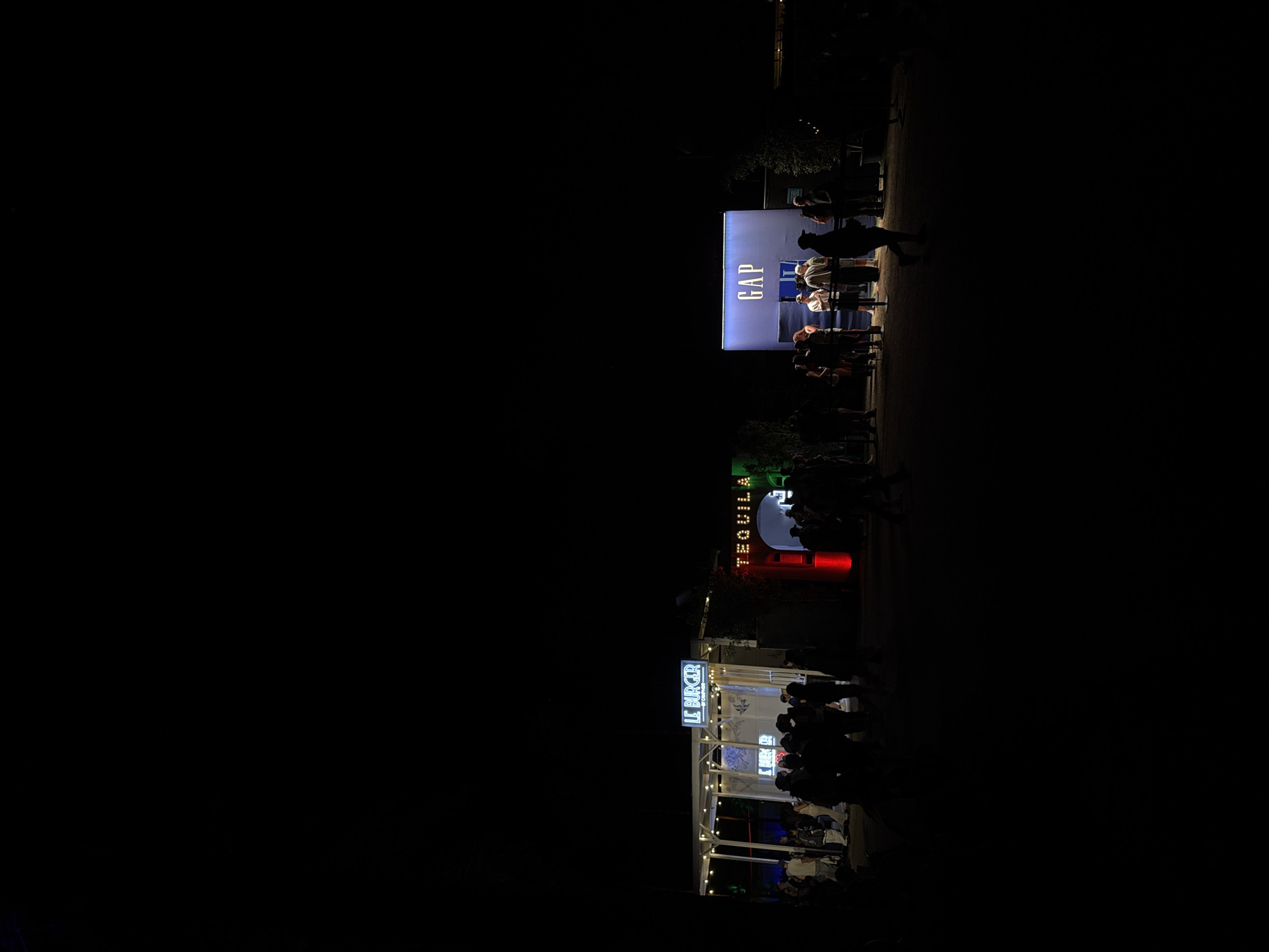 The image shows a street scene in Indio, United States of America at night.
There are three businesses or venues: a GAP store, a TEQUILA bar, and a "LE BURGER" restaurant. People are gathered at all three locations, suggesting activity and customers. The businesses are illuminated with signs and lights, contrasting with the surrounding darkness.