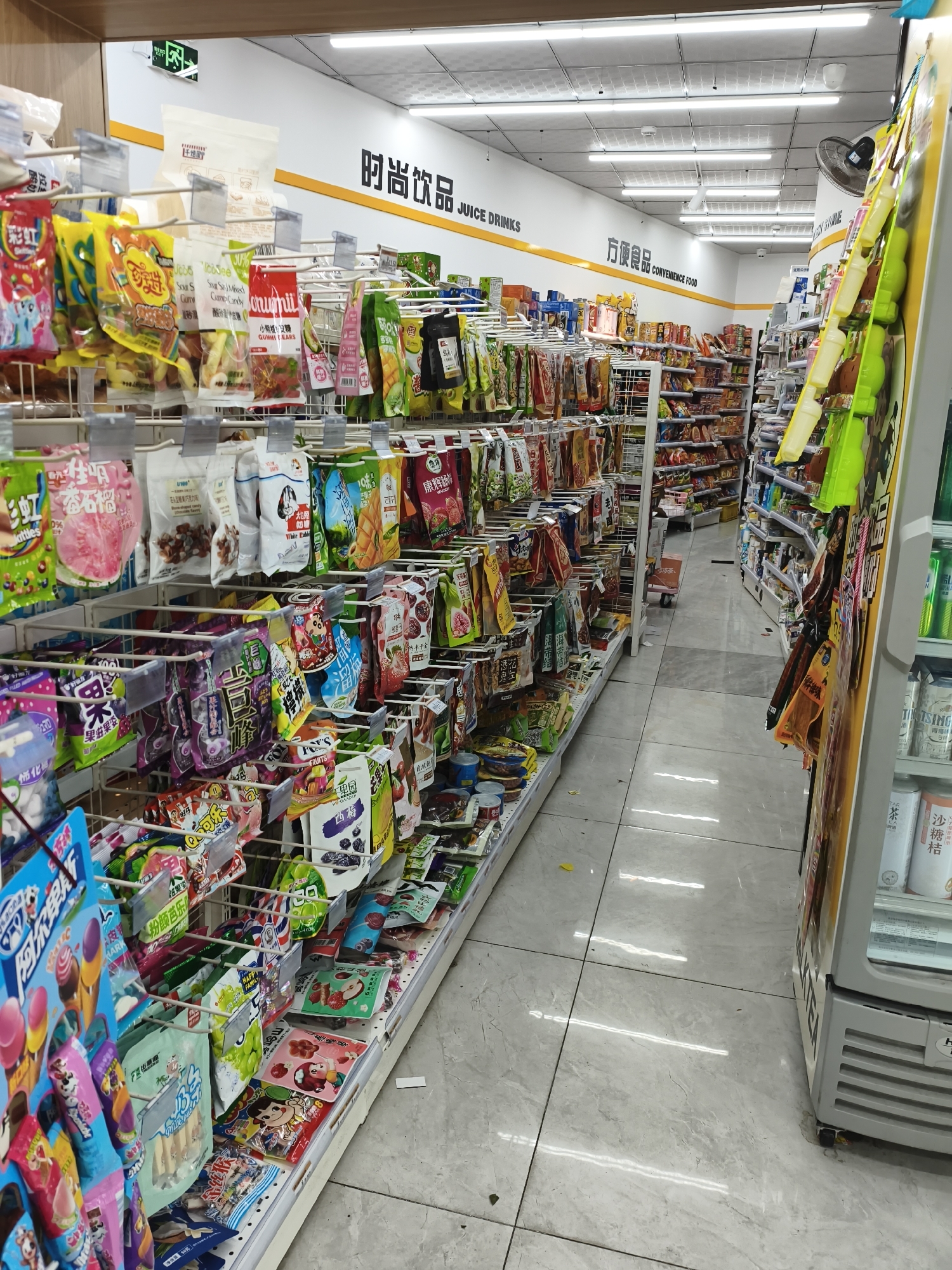 The image shows the interior of a convenience store, likely a 7-Eleven, located in Mudanjiang, China. The store features rows of shelves stocked with various snacks, candies, and beverages. A sign on the wall reads "时尚饮品 JUICE DRINKS" and "方便食品 CONVENIENCE FOOD" in Chinese and English. The floor is tiled and reflects the fluorescent lighting from the ceiling. There are no people visible in the image. The overall impression is one of a typical, well-stocked convenience store.