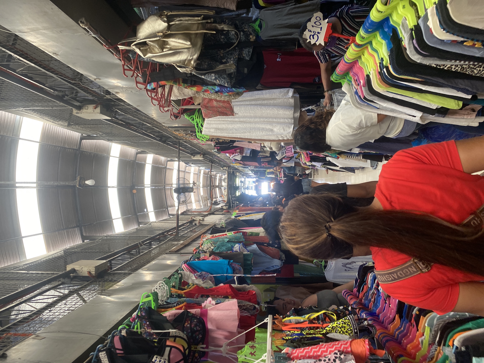 The image captures a bustling indoor market, likely a second-hand clothing store or "ukay-ukay," viewed from an elevated perspective looking down a long aisle. Several people, primarily women, are engaged in browsing and shopping for clothes.
**Subjects:** The foreground shows the back of a person with long brown hair in a ponytail, wearing a red top with a visible strap that reads "LINGDAISHU." They are looking down at racks filled with brightly colored clothing on various hangers. Other shoppers are seen further down the aisle, obscured by clothing racks. One person in a striped shirt is seen looking at clothes near a sign.
**Objects:** The market is packed with clothing items, including shirts, dresses, and other garments, displayed on metal racks and in piles. Many clothes hang on colorful plastic hangers (pink, green, blue, yellow). A gold-colored handbag is visible among the hanging items. The market infrastructure includes metal grates, beams, and corrugated metal walls/ceiling, suggesting a large, warehouse-like building. A bare light bulb hangs from the ceiling, and a ceiling fan is visible in the distance. A white electrical box is mounted on the upper structure.
**Activity or event taking place:** The primary activity is shopping or browsing for clothing. People are leaning over racks and examining items.
**Notable details:** The scene suggests a vibrant, albeit crowded, shopping environment. Bright light streams in from the side, indicating it is daytime. The general setup points to a local market.
**Visible text:** A prominent white sign displays "3 for ₱100," indicating a bundled price in Philippine Pesos. Another partially visible sign shows "10." Text resembling "LINGDAISHU" is on the strap of a red shirt, and "FASHION" appears on some tags. A red shirt has "2-1" printed on it, and some Chinese characters are visible on a green garment further down the aisle.