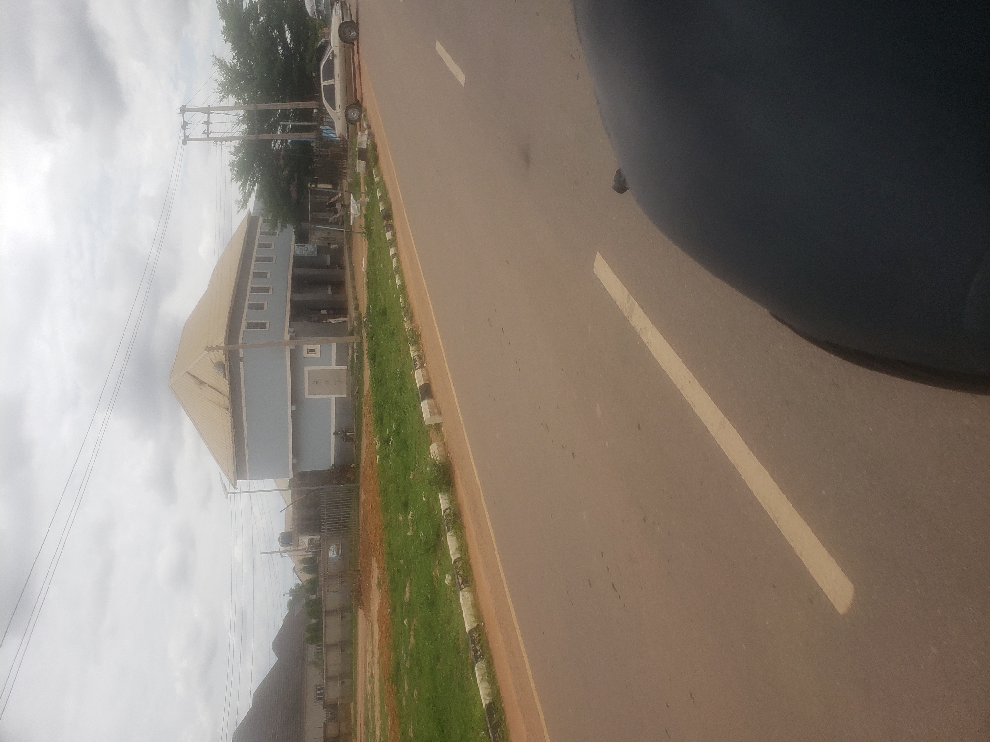 This image depicts a street scene in Rido, Nigeria, under an overcast sky. The lighting is diffused, resulting in muted colors and a lack of harsh shadows.
In the immediate foreground, the dark-colored hood and fender of a vehicle are visible on the bottom right, suggesting the photograph was taken from within or atop another vehicle. The asphalt road extends from the foreground into the mid-ground, marked with a continuous yellow line along its left edge and dashed white lines indicating lanes.
To the left of the road, a curb painted with alternating black and white sections separates the road from a dirt shoulder. Beyond the curb, a strip of green grass and patches of reddish-brown earth are visible.
In the mid-ground, a two-story building with a light blue lower facade and a golden/tan hip roof dominates the left side. The upper floor features multiple rectangular windows. The ground floor appears to have an open-fronted area or veranda supported by pillars, with a closed door visible on the far left of the ground level. A faint human figure in dark clothing is discernible standing near this door. In front of this building, a dark metal fence separates the property from the roadside. To the right of the building, a modern street light on a pole is visible.
Further to the right, a tall utility pole equipped with multiple cross-arms and electrical transformers stands prominently. A light-colored sedan (possibly silver or beige) is parked on the road's shoulder, facing right, near the utility pole. A second human figure, dressed in dark attire, is standing near the base of this utility pole.
In the background, power lines stretch across the upper portion of the sky. Additional residential structures with dark roofs are partially visible to the left and behind the main blue building. Trees with dense green foliage are present to the right of the utility pole, partially obscuring the distant background. A water tank on a raised stand is faintly visible behind the fence in the mid-ground. The overall scene suggests a suburban or developing area.