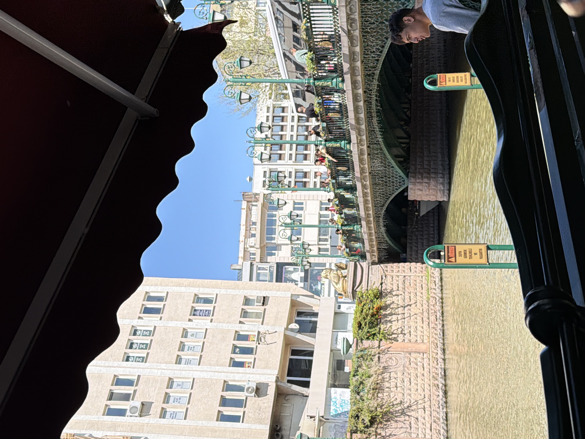 The image captures a vibrant daytime scene in Eskisehir, Turkey, likely along the Porsuk River. The perspective is from beneath a dark red, scalloped awning, possibly from a cafe or restaurant, looking out over the water and a bustling city environment.
In the foreground, the dark red awning forms a significant part of the left and top edges. A young man with short, dark curly hair, wearing a light grey t-shirt, is visible on the right, looking down towards the murky, light greenish-brown water below. He appears to be leaning against a dark metal railing that runs along the edge of the viewing point.
Below him and extending across the frame are two yellow and green warning signs attached to green metal loops by the water's edge. The text on these signs, written in Turkish, reads "DİKKAT! SUYA GİRMEK TEHLİKELİ VE YASAKTIR." which translates to "ATTENTION! ENTERING THE WATER IS DANGEROUS AND FORBIDDEN."
Across the water, a decorative green metal bridge or walkway is visible, lined with ornate lampposts featuring multiple lights and flower boxes. Several people can be seen standing and strolling on this structure. Beyond the bridge, modern buildings with numerous windows and some air conditioning units line the riverbank under a clear blue sky. A golden lion statue is perched on the bank between the bridge and the buildings. Some windows on the buildings display signs, including a partial phone number "0542...". The general atmosphere is bright and sunny, indicating good weather.