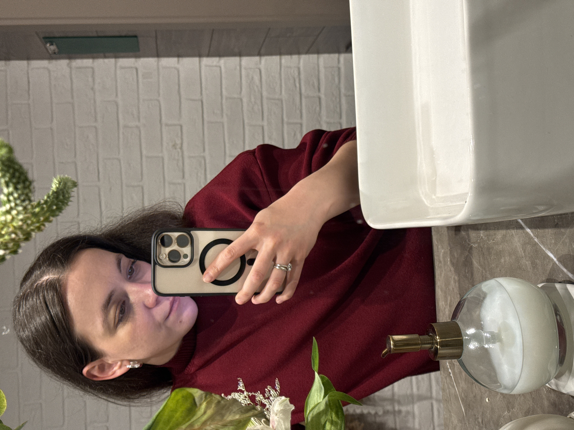 A woman with dark hair is taking a selfie in a bathroom. She is wearing a maroon sweater and holding a smartphone with a beige case. A white sink is partially visible in the foreground, and a soap dispenser sits on the countertop. The background features a white brick wall. The location is likely a bathroom in the city of Chaikovsky, Russia.