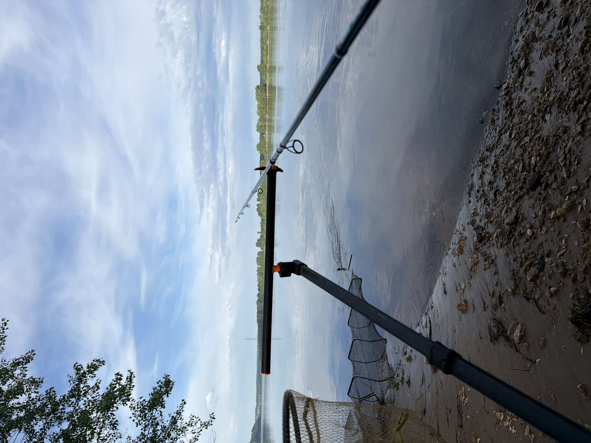 The image shows a fishing setup at the edge of a body of water, likely a river or lake. A fishing rod is placed on a rod rest, and a landing net is positioned next to the water. The shoreline is visible with some vegetation. In the background, across the water, a treeline can be seen. The sky is partly cloudy. While the query requests the location in the city of Noviy, Russia, there is nothing in the image to explicitly confirm or deny this location. The location could be deduced if there was some landmark, a specific building, or a recognizable feature present in the image. Without this information, it's impossible to confirm the location.