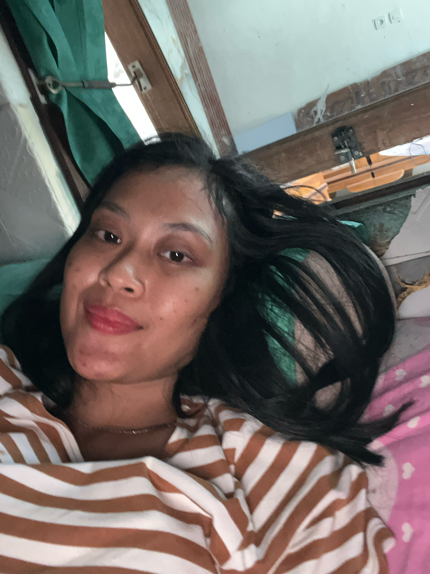A woman lies in bed, her head propped up and her body angled towards the camera. She has dark hair, which is mostly spread out around her head, and her face is turned slightly to her left. She is wearing a brown and white striped shirt. Her right arm is visible, bent at the elbow. Behind her, a green curtain hangs from a hook on a wooden frame, and to her right, a pink blanket with white heart shapes is visible. The background also shows elements of a room, including wooden structures and a white ceiling with some markings.