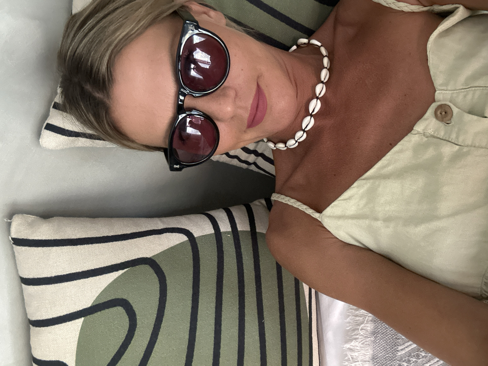 A fair-skinned woman with blonde hair and sunglasses is reclining on a couch. She is wearing a light-colored top and a shell necklace. The couch has cushions with green and black stripes. The location is implied to be North Kuta, Indonesia.
