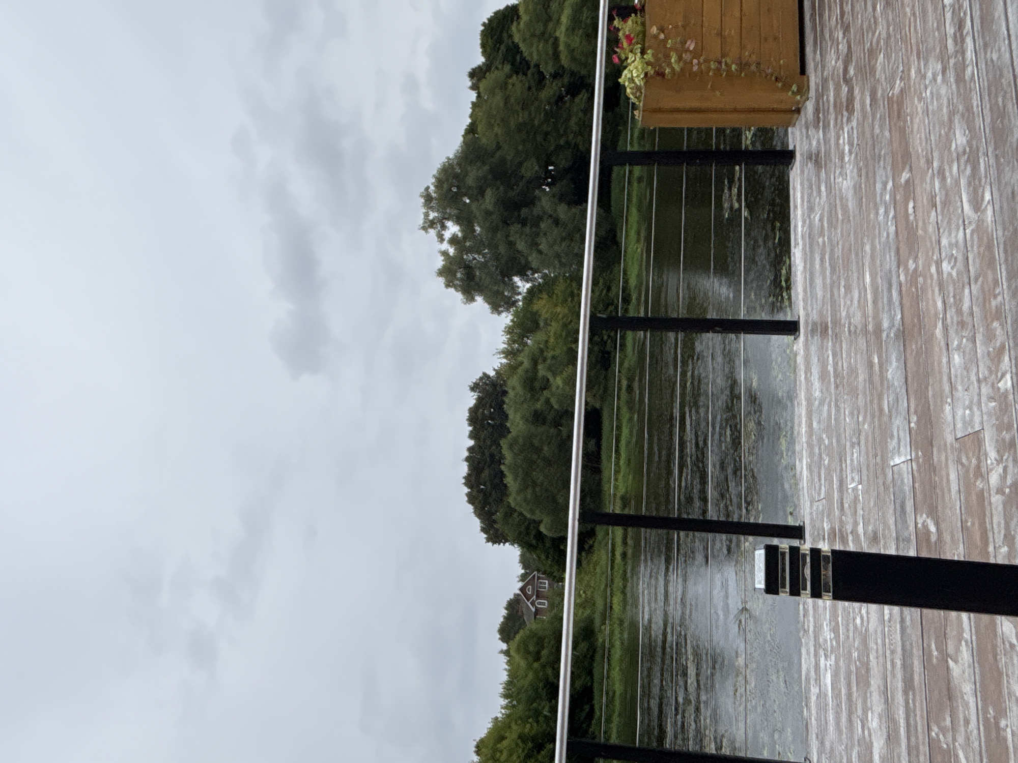 The image depicts a cloudy, overcast day scene from a wooden pier overlooking a calm body of water. In the background, a lush green treeline borders the water, with a small, reddish-brown house visible amongst the foliage. The pier has a metal railing with vertical black posts and horizontal silver bars. A black bollard-style light fixture stands on the pier in the foreground, near a wooden planter box filled with greenery and pink flowers. The wooden planks of the pier appear weathered and slightly damp. The overall atmosphere is serene and natural, suggesting a peaceful riverside location.