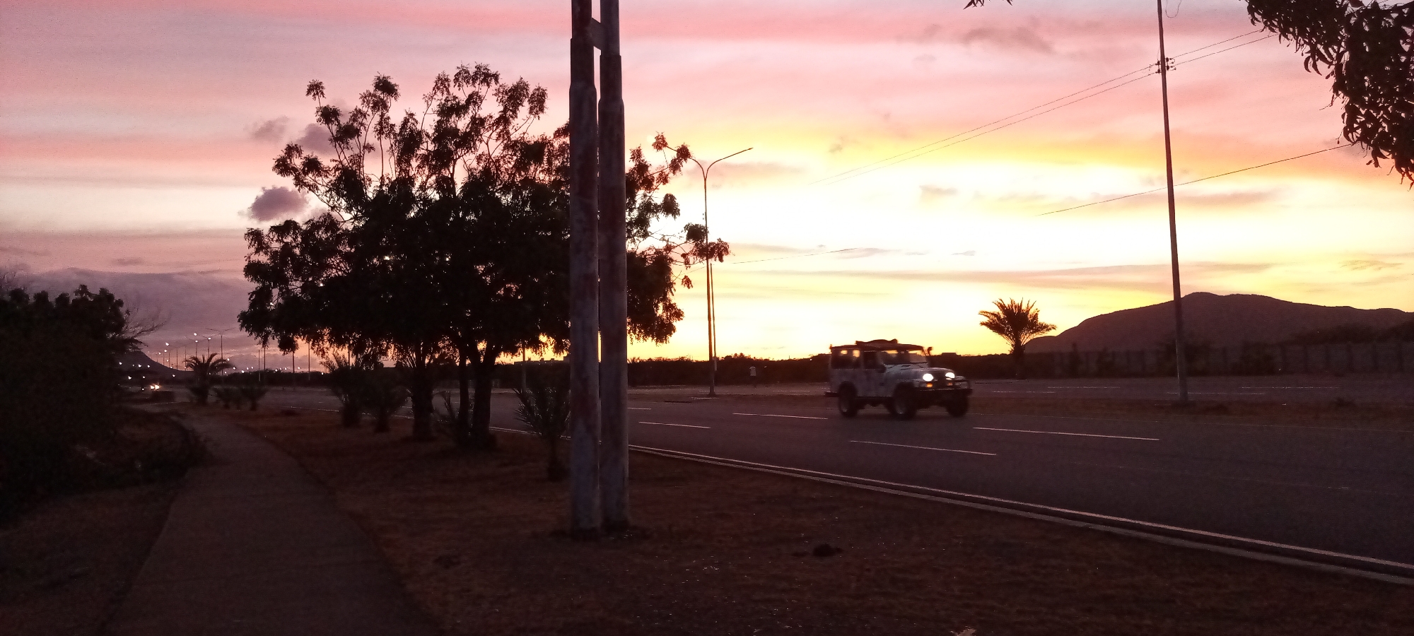 The image captures a sunset scene on a road in an urban or suburban environment. The sky is filled with colors ranging from pink to orange and yellow. Along the left side of the road is a sidewalk, with trees of varying sizes lining the roadside. On the right side of the road is a mountain. Streetlights are present alongside the road. A jeep is visible on the road, moving towards the viewer. The location appears to be a coastal city, although the specific location (city and state) is unknown.