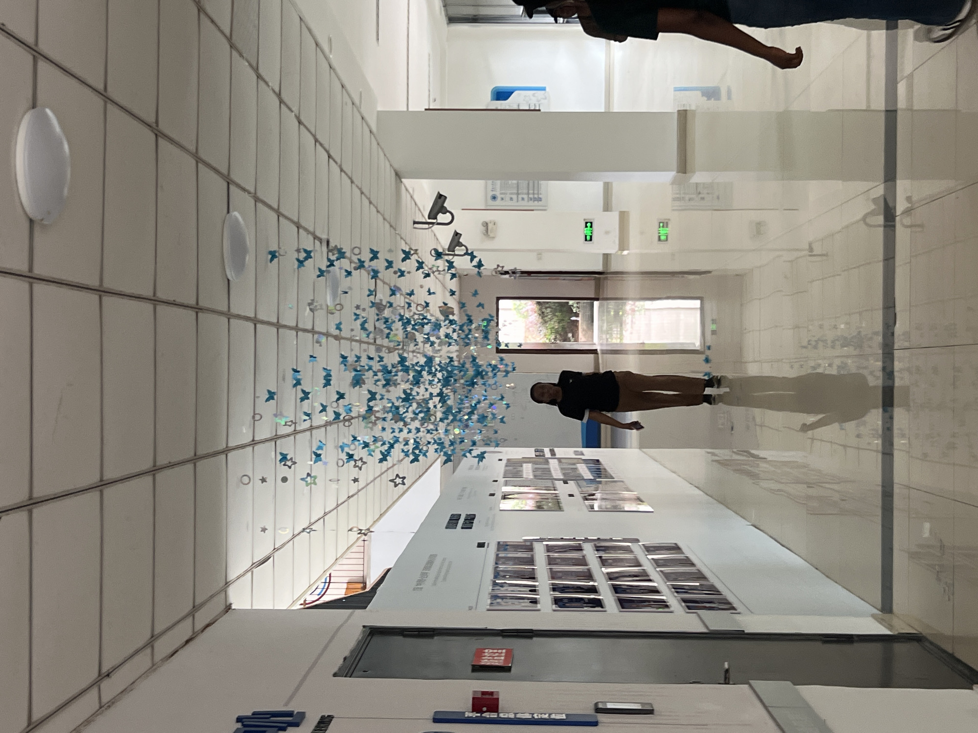 The image shows an indoor hallway with a tiled floor and a dropped ceiling. On the left wall, there is a series of framed photographs or art pieces. A woman in a black t-shirt and brown pants is walking down the hallway, casting a reflection on the polished floor. Suspended from the ceiling is a decorative installation of blue and silver butterfly-shaped ornaments. Two security cameras are visible on the right side of the hallway. Green illuminated exit signs are present on the right wall. The hallway appears to be a public space within a building, possibly an educational institution or a gallery. The scene is well-lit by overhead fluorescent lights and possibly natural light from a distant opening. The overall impression is that of a modern, clean, and possibly artistic interior space.