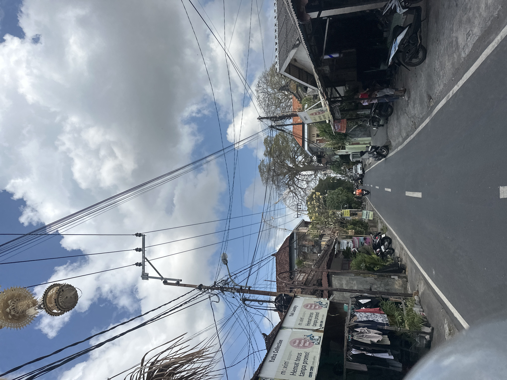 The image depicts a street scene in Denpasar, Indonesia, characterized by a narrow asphalt road bordered by buildings and a dense network of overhead electrical wires. On the left side of the road, a clothing rack displays various garments. Adjacent to it, a sign for "Yutha Collection" and "Yuki Laundry" with promotional text is visible. Further down the street, several motorcycles are parked. The buildings are of traditional Balinese architecture, with tiled roofs. On the right side of the road, shops with open fronts are present, and more motorcycles are parked. Two individuals, possibly a parent and child, are standing on the sidewalk in front of one of the shops. A few more motorcycles are seen on the road further ahead, and a distant figure on a motorcycle is visible. The sky is partly cloudy. A large tree with bare branches stands prominently in the background.