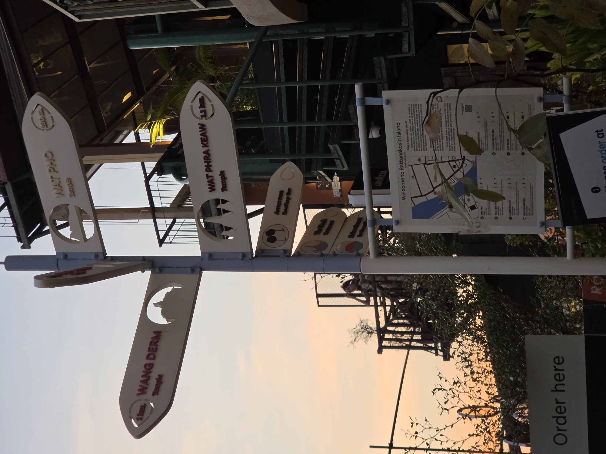 The image shows a signpost in Bangkok, Thailand, specifically pointing to different landmarks on Rattanakosin Island. The signpost features directional arrows indicating distances to locations like Wat Pho (230 m), Wang Derm Temple (1 km), and Wat Phra Keaw Temple (1.1 km). There are also signs for Amorosa Rooftop Bar and other places. A map entitled "Welcome to Rattanakosin Island" is attached to the pole. In the background, the sky is a soft orange at sunset. The sign is located near an outdoor area, as indicated by foliage and partial structures like scaffolding and a staircase.