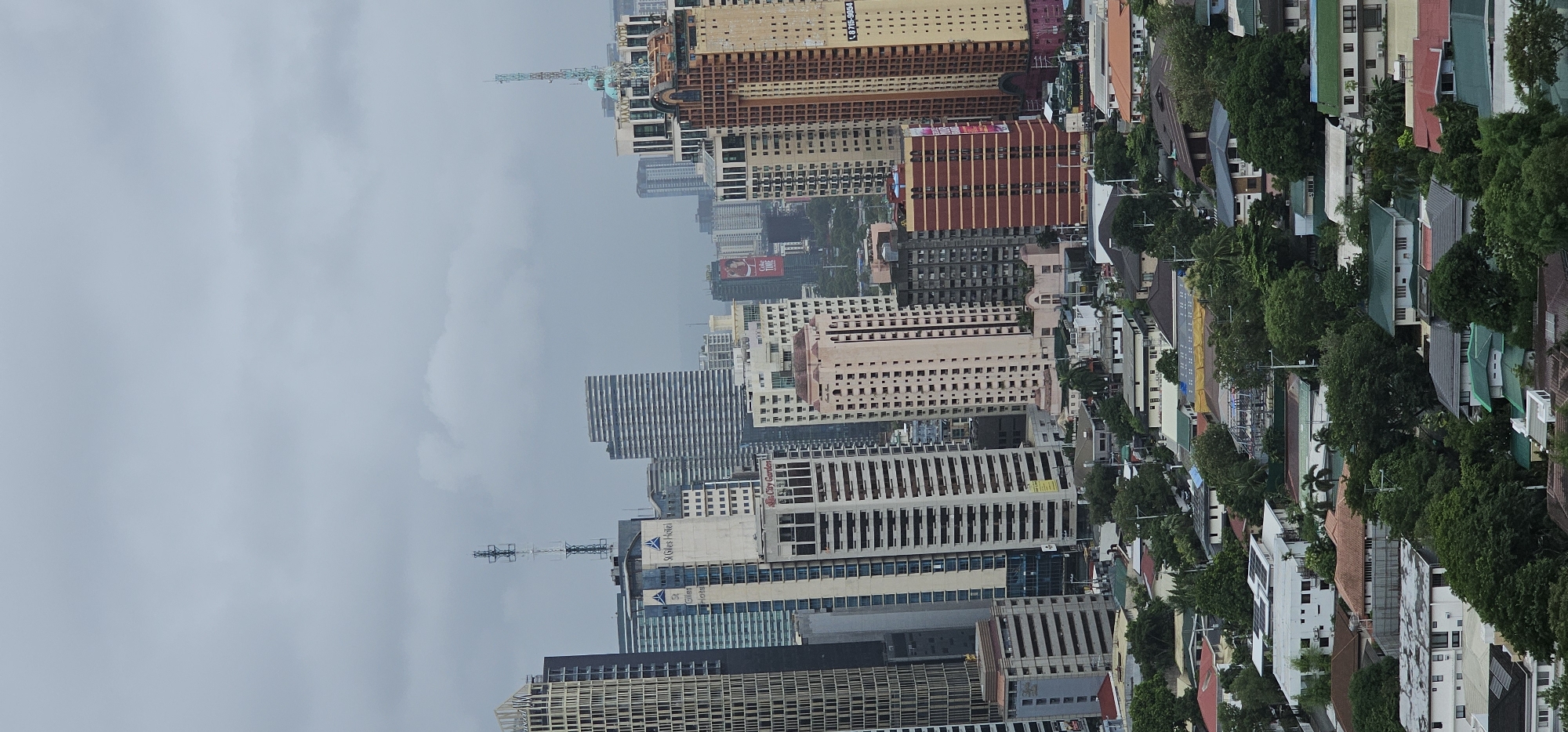 The image shows a dense urban cityscape, likely Makati City, Philippines, on a cloudy day. The foreground consists of a suburban neighborhood with numerous houses, many with tiled roofs in red, brown, and green. These houses are surrounded by lush green trees. Behind this residential area, the scene transitions to a dense collection of high-rise buildings, predominantly skyscrapers.
These buildings vary in architectural style and color, with prominent examples including a tall, slender building with a checkered facade on the left, a cream-colored building with "St. Giles Hotel" signage, and a striking orange and brown building on the right with a communication tower on its roof. Other notable structures include a building with a Coca-Cola advertisement visible, and a cluster of residential-style buildings integrated with commercial ones. Several communication towers and antennae are visible atop some of the buildings. The overall impression is of a sprawling metropolitan area where residential areas coexist with commercial and business districts. There are no visible people in the image.