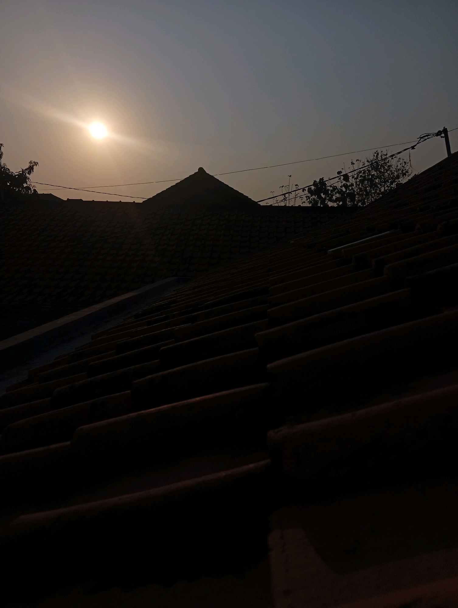 The image depicts a sunset over a residential area in Kebandingan, Indonesia. The sun, a bright orb with visible rays, is positioned in the upper left quadrant of the sky, casting a warm glow. The sky is a gradient of colors, transitioning from a pale yellow near the sun to a dusky blue at the top. In the foreground, the tiled rooftops of houses dominate the scene, rendered in silhouette against the fading light. The texture of the tiles is evident, with rows of curved shapes creating a sense of depth and pattern. A lone utility pole with wires is visible on the right side of the frame, silhouetted against the sky. The overall atmosphere is one of quiet dusk in a suburban or semi-urban environment.