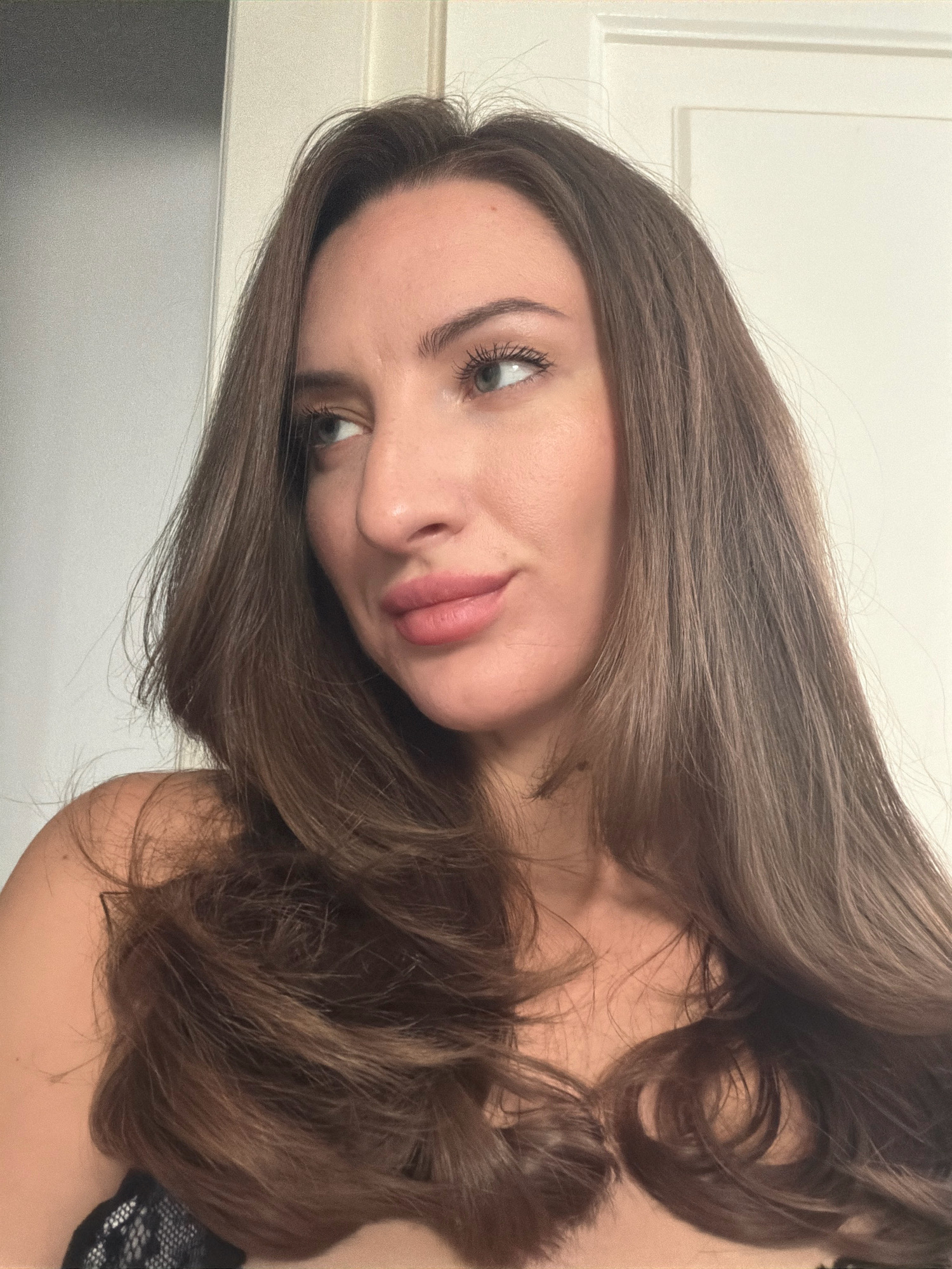 A close-up, indoor portrait shows a woman with long, wavy brown hair and light blue eyes. She is looking to her left, with her head tilted slightly. Her lips are parted and appear to be slightly pink. She is wearing what appears to be a black lace garment on her upper left shoulder. The background consists of a plain white door and a grey wall. The image does not contain information that places it in Sharm El Sheikh, Egypt.