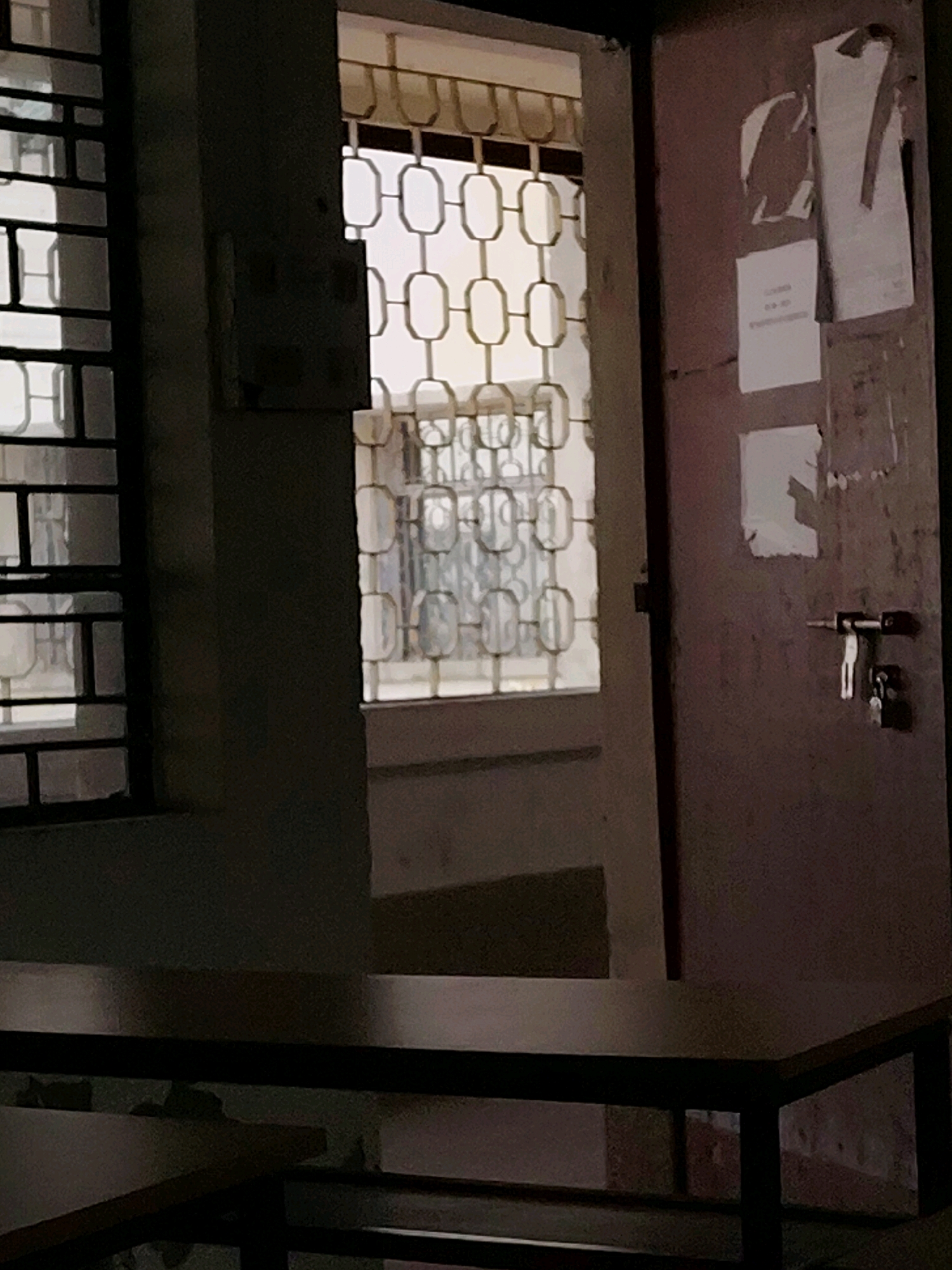 The image depicts an interior scene, possibly an office or classroom, in Jirania, India. A partially open door on the right reveals a dark pink door with a lock and key mechanism. The door is adorned with several torn pieces of paper, some appearing to be notices or posters. To the left of the door, a window with a decorative metal grille shows a second window behind it, also with a similar grille. The area in front of the window is dimly lit, with a table in the foreground. The overall impression is one of an older, somewhat unkempt indoor space.