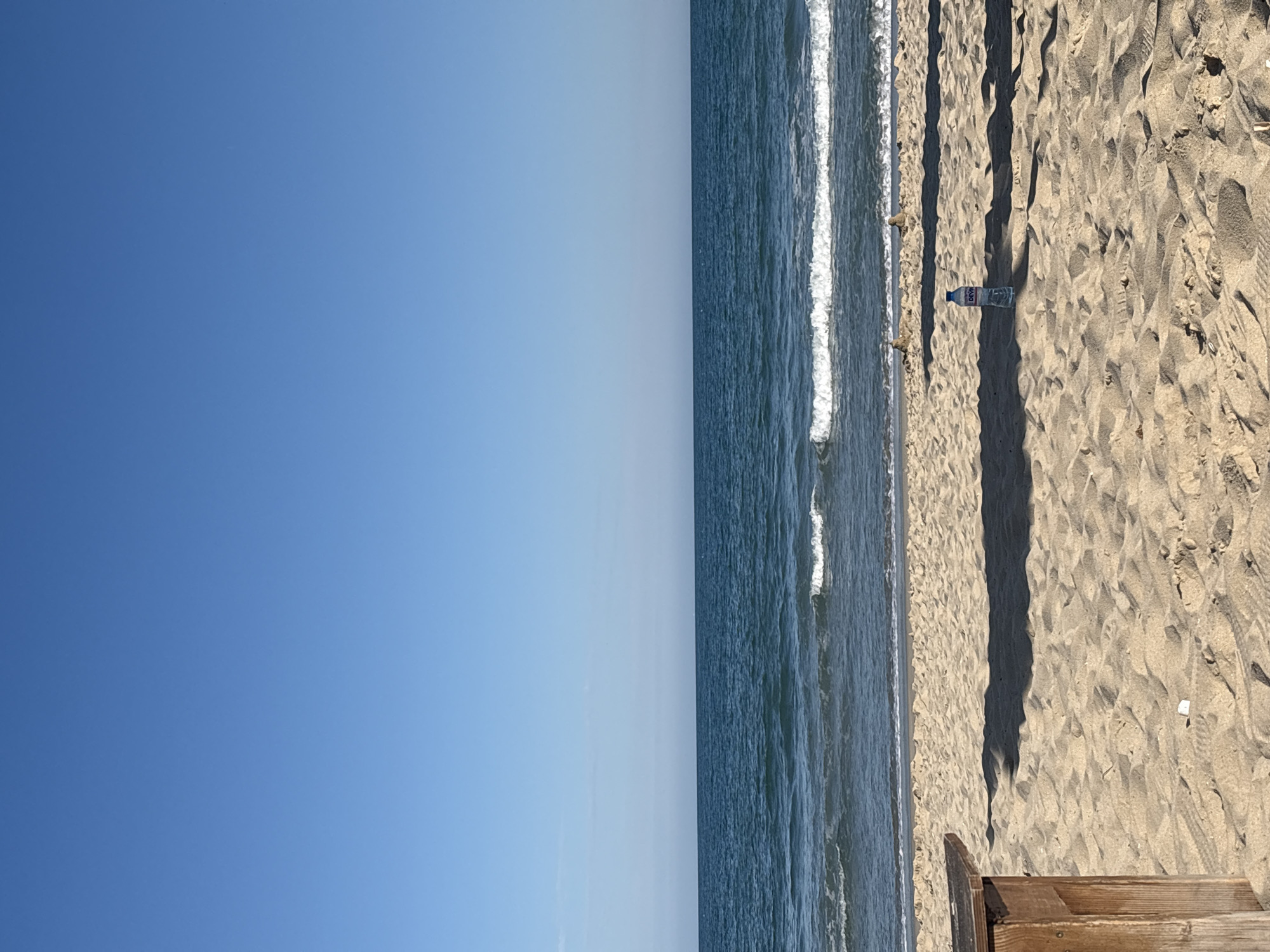 The image depicts a beach scene, likely in Varna, Bulgaria, given the coastline. To the left is a clear blue sky. To the right is a sandy beach with visible footprints and shadows, suggesting the sun is at an angle. The sea is between the sky and the beach, with small waves visible. There is a water bottle resting on the sand. A wooden structure is at the lower right corner. There are no people visible, but there are footprints in the sand.