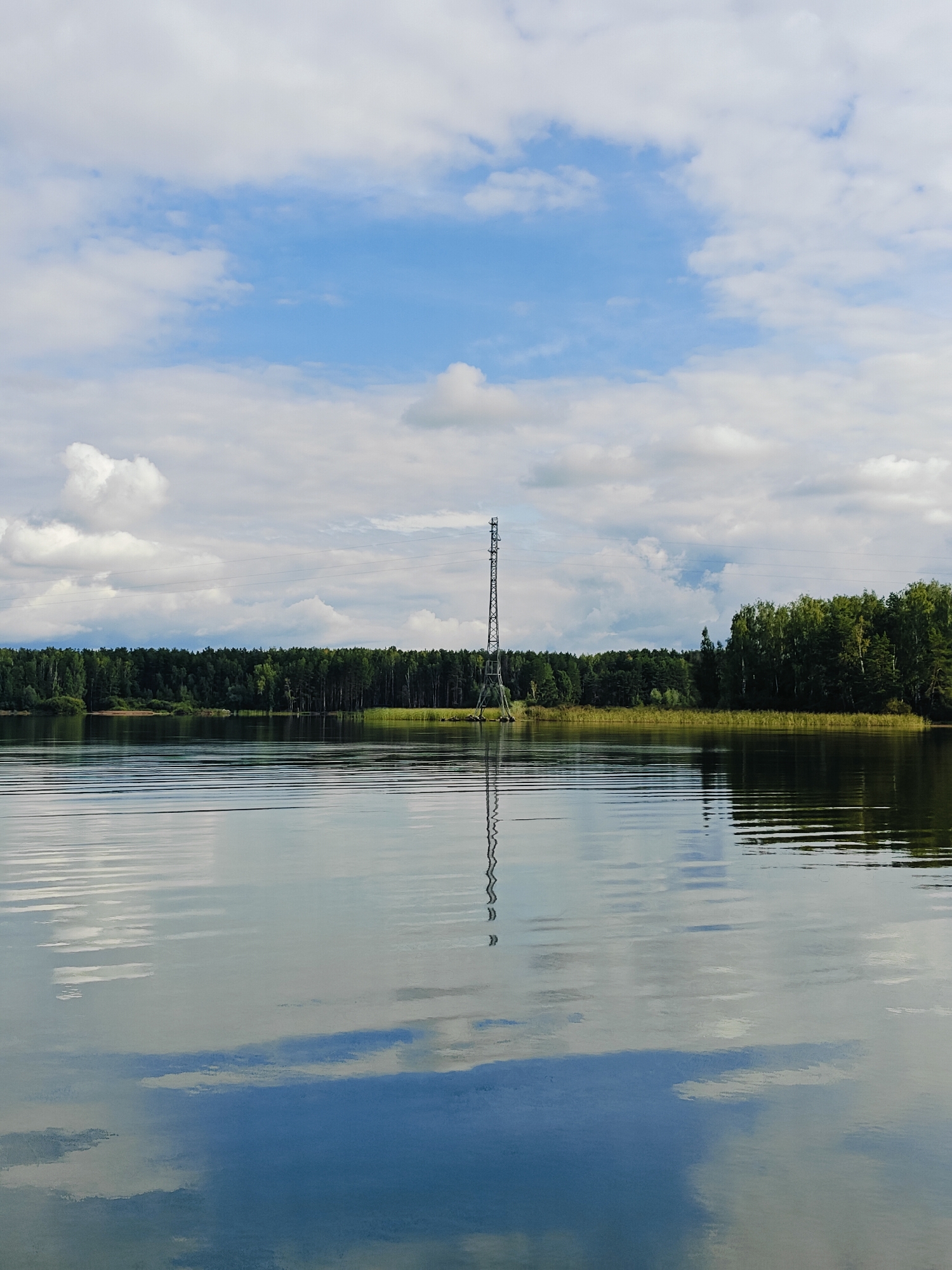 The image shows a serene lake with a dense forest lining the distant shore. A tall, metallic tower stands prominently on the left side of the lake, its reflection visible in the calm water. The sky above is a mix of blue and white clouds. The scene appears to be located in the city of Chaykovsky, Russia, a place known for its natural beauty and proximity to water bodies.