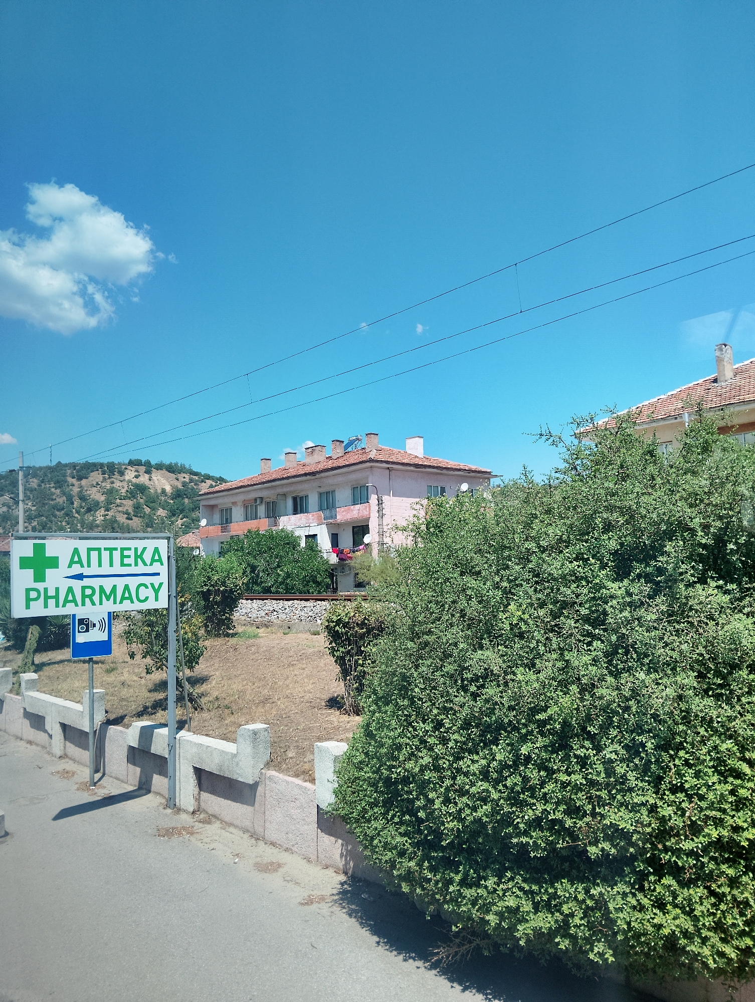 The image shows a pharmacy sign with a green cross and the words "АПТЕКА PHARMACY" written in English and Cyrillic, with an arrow pointing to the right, in Kresna, Bulgaria. The sign is mounted on a pole next to a low concrete wall. Behind the wall is a street and then a dense green bush. In the background, there are two light-colored buildings with red tile roofs, and a hill covered with dry vegetation. The sky is clear blue with a few white clouds. There are power lines stretched across the upper part of the image.