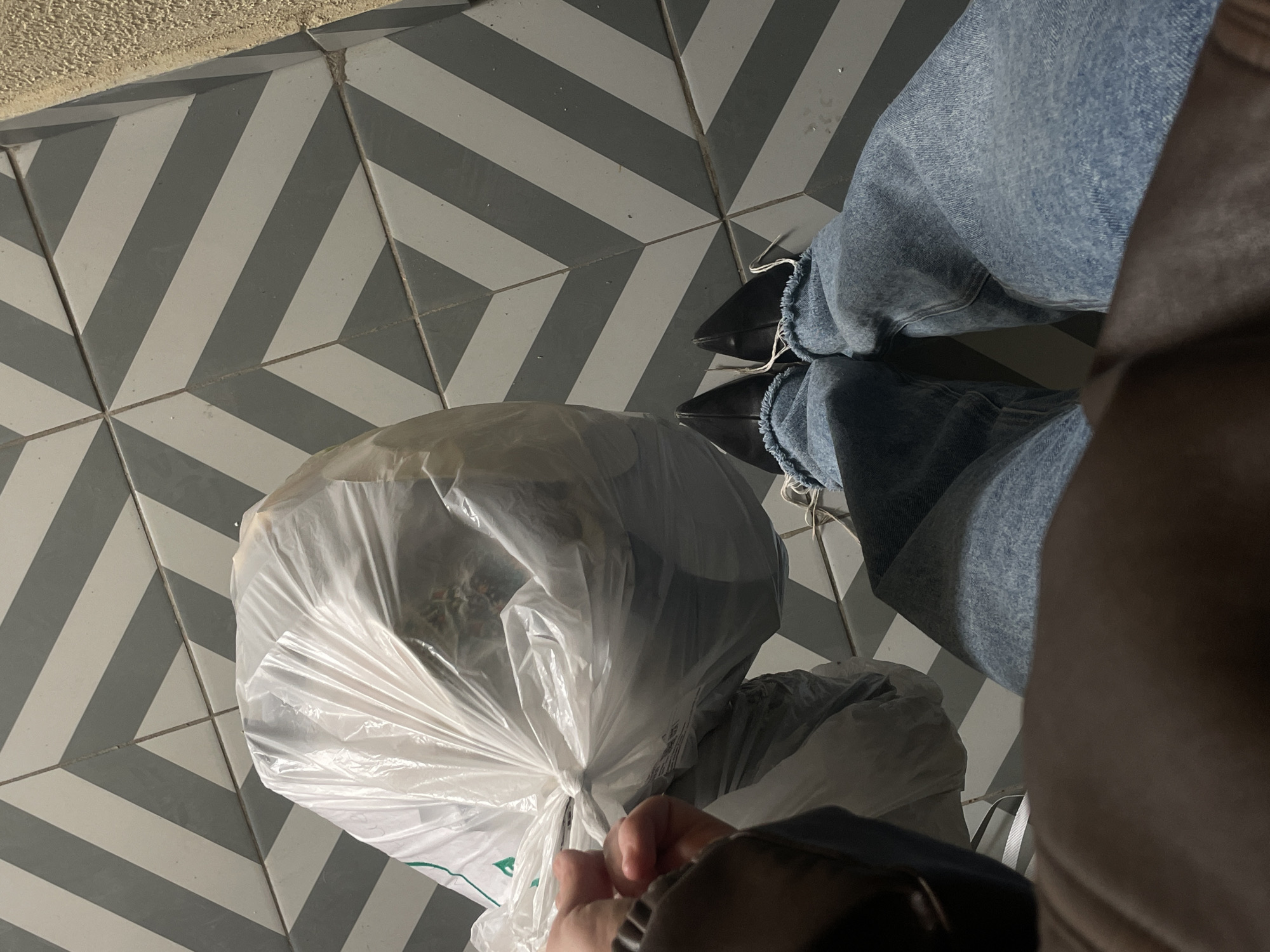 A high-angle shot depicts a person wearing light-blue, frayed jeans and black shoes, standing on a grey and white chevron-patterned tiled floor. The person is holding a partially filled, white, plastic bag closed. The scene features a brown, textured surface in the upper-left corner and the edge of a dark-brown jacket in the lower-right corner. The location of the photo is unknown. The city of Moscow, Russia, is not discernible in the image.