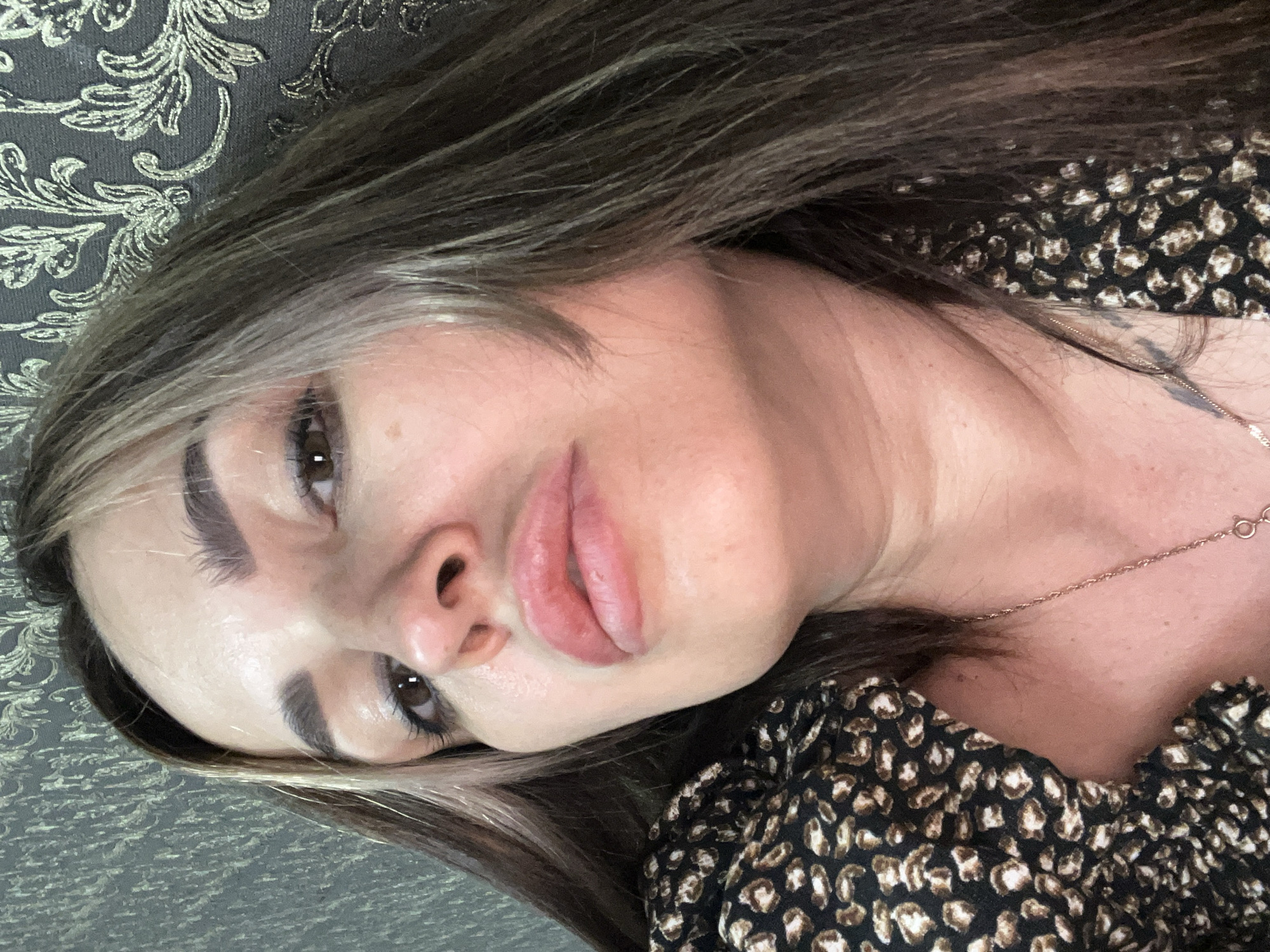 An overhead, close-up selfie of a woman with brown hair, dark eyebrows, and brown eyes, is angled towards the camera. She is wearing a black dress with a tan and white animal print pattern and a gold necklace. Her skin appears smooth and fair. The background is a wall with a textured damask pattern in a dark gray and gold color. The lighting is soft, casting a warm glow on her face.