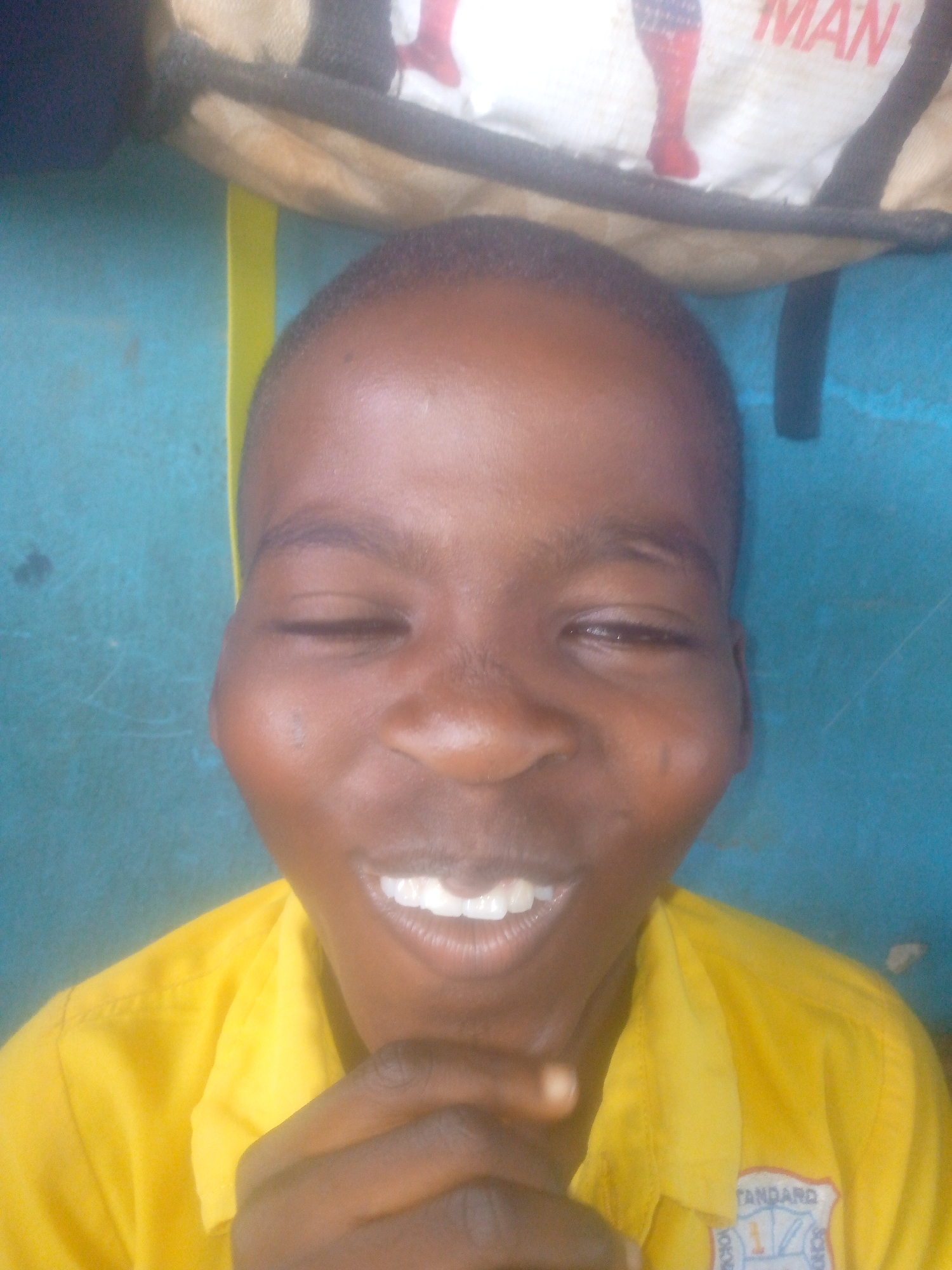 A young African male with dark skin and short hair is shown in a close-up, slightly off-center shot. The subject is wearing a yellow collared shirt with the words "STANDARD" and "SCHOOL" visible on an emblem. The subject is smiling with closed eyes and has his hands clasped together under his chin. Behind the subject is a blue wall and a partially visible bag with a cartoon-like image of "Spiderman" and the word "MAN." The photo may have been taken in Ikorodu, Nigeria.