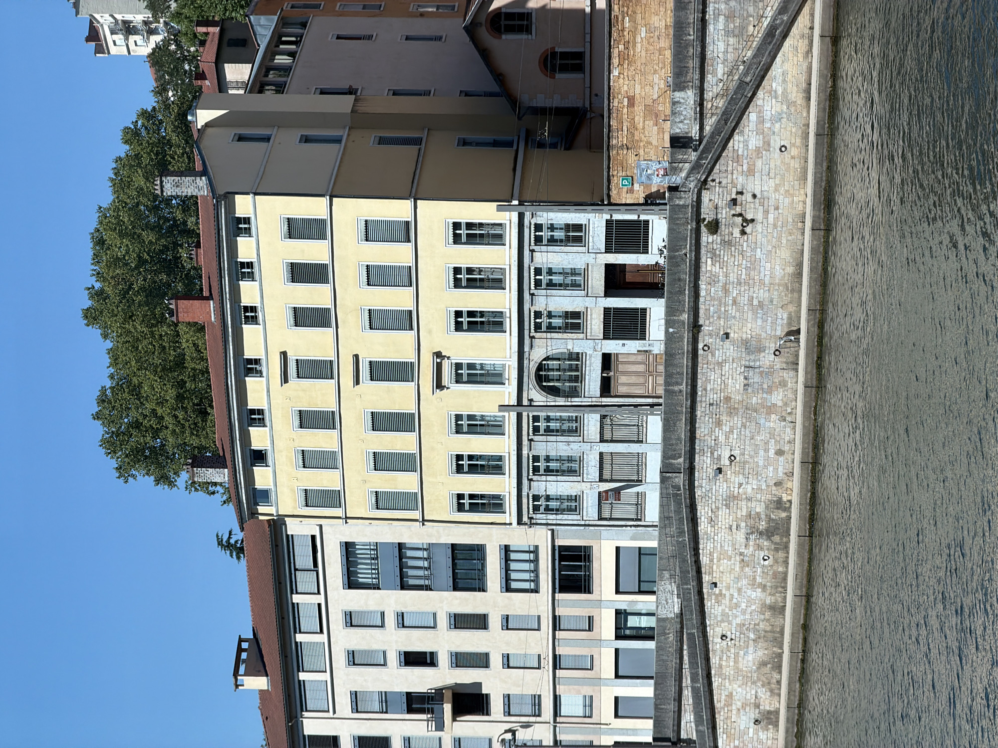 The image shows a view of buildings and a body of water, likely a river, along a waterfront. The buildings are multi-story structures with a variety of architectural styles. One prominent building is painted a pale yellow and has numerous windows with white shutters. Next to it, another building features grey stone facades and elaborate window grilles. Further back, a building with a red-tiled roof and a brick chimney is visible, partially obscured by lush green trees. In the foreground, a paved walkway runs alongside the water, lined with stone and featuring some metal rings embedded in the ground. The water has a textured surface, suggesting it is flowing. The sky is clear and blue, indicating a sunny day. The scene appears to be a cityscape, possibly in Lyon, France, given the architectural styles and the presence of a river. The image is taken from an elevated perspective, looking down on the scene. There is no visible text.