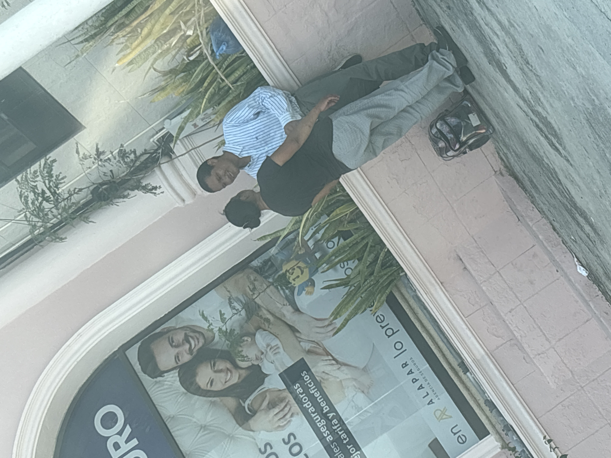 Two men are standing in front of a storefront at an oblique angle. One man is wearing a light blue and white striped shirt and dark pants, and the other is wearing a dark shirt and light-colored pants. The storefront window displays an advertisement featuring a smiling family and text in Spanish, including "RO" and "en ALAPAR lo preve AGENCIA DE SEGUROS." Plants are visible along the building facade. Based on the text in the advertisement, the location is likely in Tampico, Mexico.
