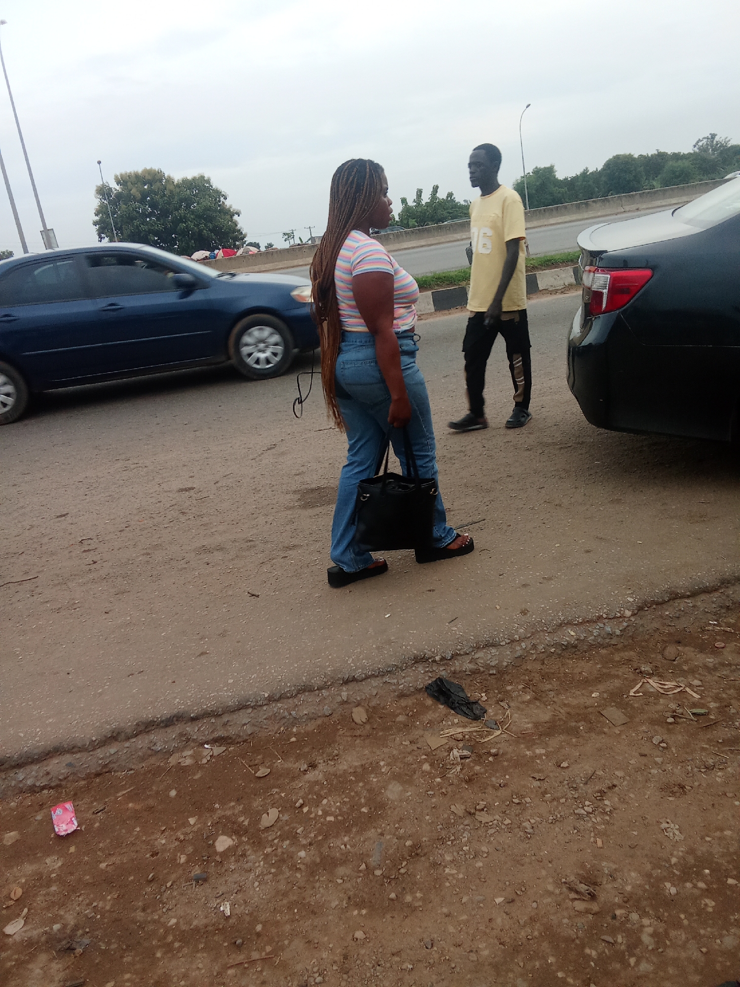 Two individuals stand on a dusty roadside, their interaction obscured by the image's focus on their full bodies and the surrounding environment. A woman, facing away from the camera, wears a horizontally striped t-shirt and denim jeans, carrying a black handbag. Her hair is styled in long braids. A man, also with his back to the viewer, is dressed in a yellow t-shirt with a graphic and dark, ripped pants. They appear to be waiting or conversing near a roadside.
The scene is set on a street in Kagini, Nigeria, characterized by its unpaved shoulder and a busy road with passing cars. A blue sedan is visible in the left lane, its wheels blurred by motion. Further down the road, a dark-colored car is parked, its taillights illuminated. The background reveals a lush green environment with trees and a cloudy sky, suggesting it might be daytime with overcast weather. The road itself is a mix of asphalt and dirt, with debris scattered along the shoulder, including plastic bags and other discarded items. The overall impression is a snapshot of everyday life on a roadside in Nigeria, possibly indicating a stop or a moment of pause during travel.