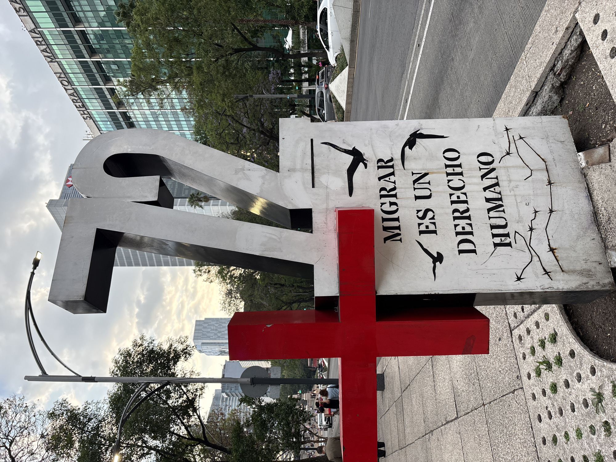This is a photograph taken at an eye-level perspective, showing a monument dedicated to migrants in Mexico City, Mexico. The monument features a large, metallic number "72" beside a white sign bearing the message "MIGRAR ES UN DERECHO HUMANO" (Migrating is a human right), along with images of birds and barbed wire. A red cross is incorporated into the monument’s base. The monument is located on a sidewalk paved with gray concrete tiles, adjacent to a curb. In the background, modern architecture is visible, including tall office buildings such as the Sheraton Maria Isabel Hotel, some with cloudy skies as the backdrop. People can be seen standing nearby. Streetlights flank the monument.