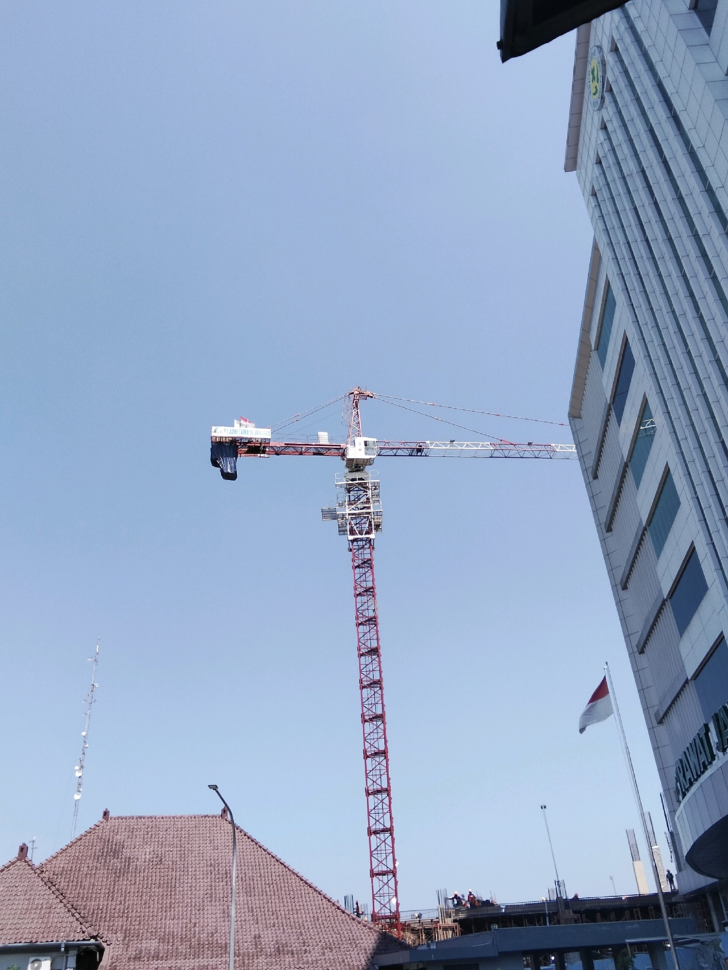 The image depicts a construction site in Magelang, Indonesia, under a clear, pale blue sky. A tall, red and white construction crane dominates the center of the frame, its jib extending horizontally. A dark blue tarp is draped over a portion of the jib. Two Indonesian flags are visible on the jib of the crane. To the right, a modern, multi-story building with a white, tiled facade and dark windows is partially visible, its angled perspective drawing the eye upwards. The building has signage that includes the words "RAWA" in large, dark letters. In the foreground, to the left, a building with a brown, tiled roof and visible rafters is present. A street lamp stands near the center of the image. Further in the background, a lattice-like telecommunications tower can be seen. Several construction workers in hard hats are visible on scaffolding below the level of the crane's jib. A small, white flag on a flagpole is positioned near the modern building.