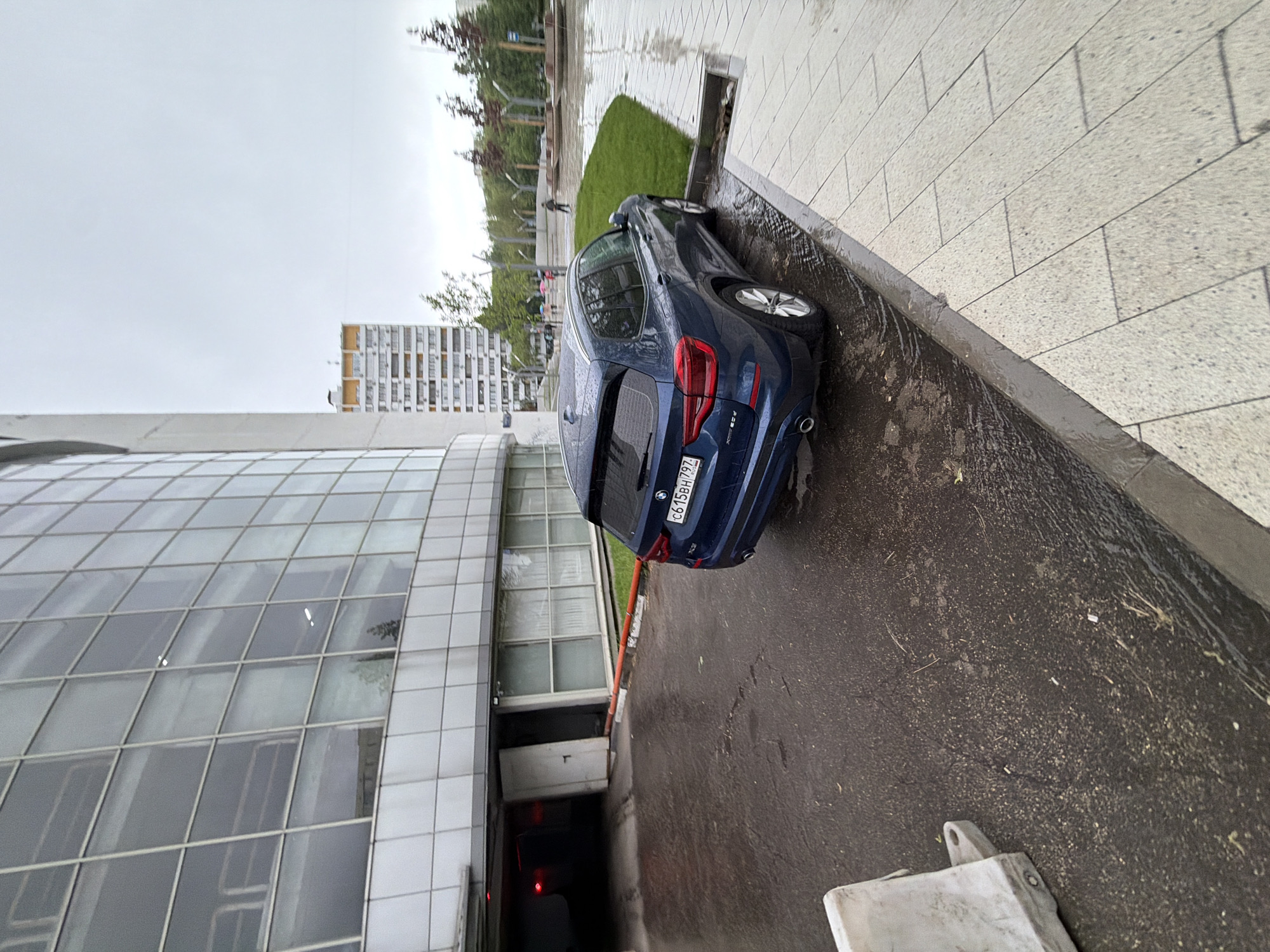 The image shows a blue BMW parked on a wet street next to a modern building with a curved, grid-like facade in Moscow, Russia. The car is parked near the entrance to an underground parking garage. A tall, apartment building is visible in the background. The weather appears overcast and rainy. A small patch of grass and a sidewalk are also visible. Pedestrians are visible in the distance.