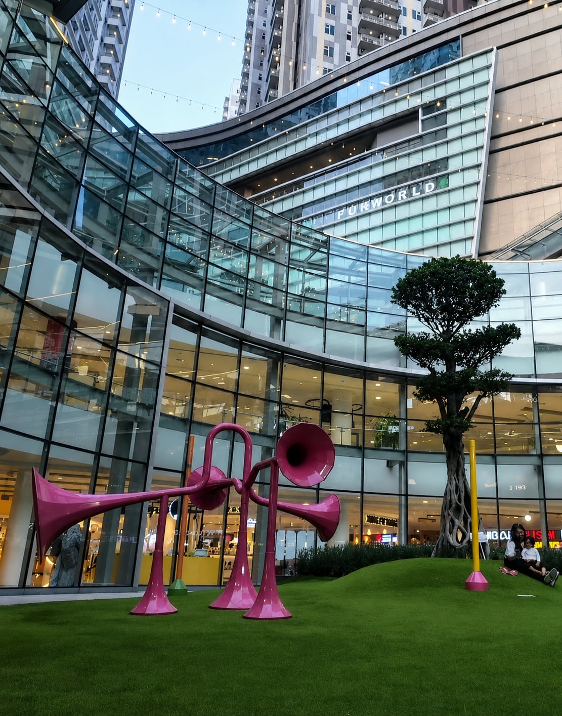 In the city of Medan, Indonesia, at the Sun Plaza mall, an outdoor area features a grassy, undulating landscape. A sculpture resembling oversized pink trumpets stands prominently on the lawn. To the right, a lone tree with gnarled roots is visible, adjacent to a yellow pole. A woman and a child are seated on the grass near the tree, appearing relaxed. The background is dominated by the modern architecture of the mall, characterized by extensive glass facades and multi-story buildings. String lights are visible, adding to the ambiance of the outdoor space.