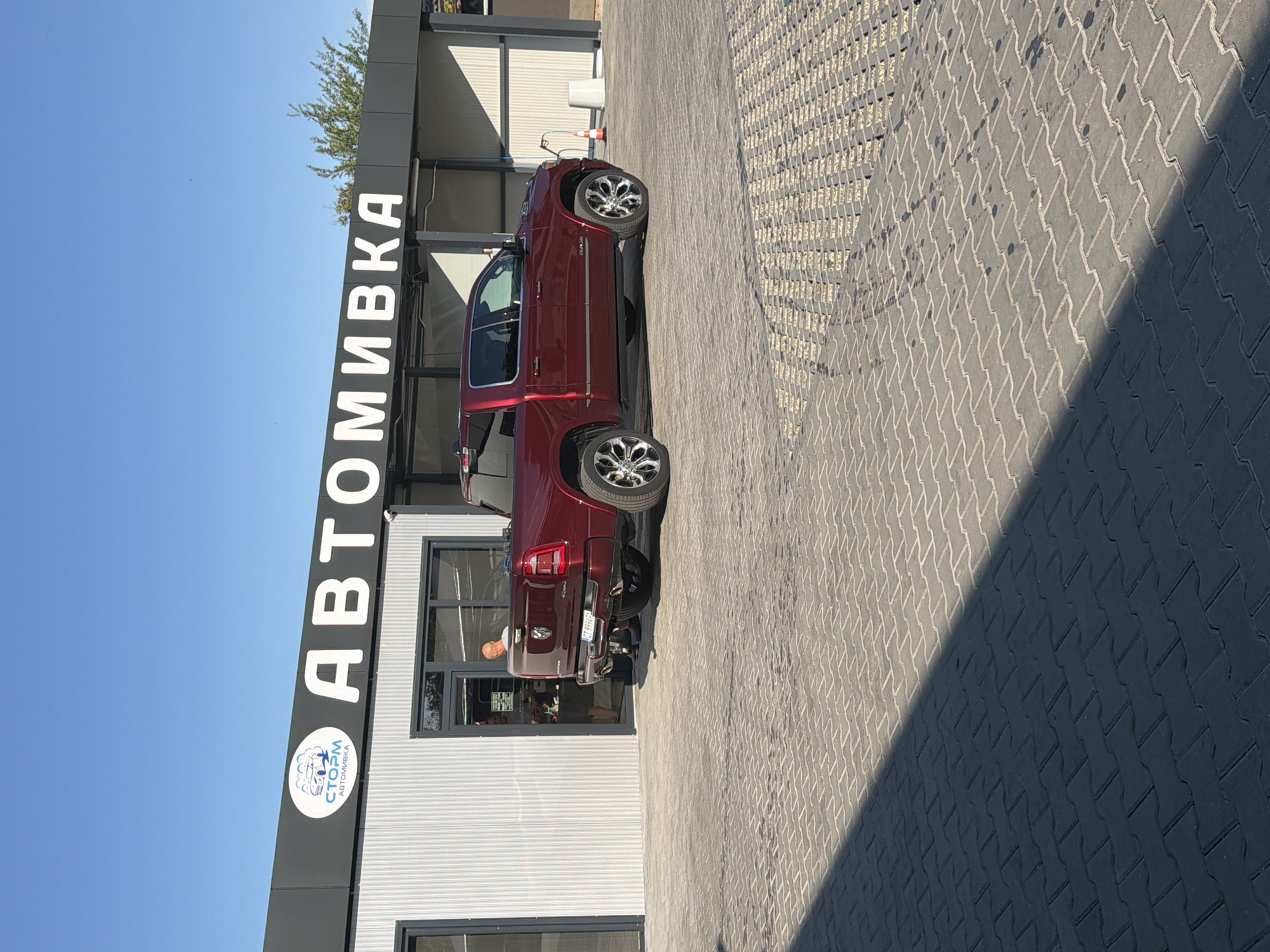 A red Ram 1500 pickup truck is parked outside a car wash facility. The car wash building has a dark gray facade with white lettering that reads "АВТОМИВКА" (which translates to "Car Wash" in Russian). Above the lettering, a blue and white logo features a stylized car and bubbles, along with the word "СТОЯМ" and "АВТОМИВКА". The truck is facing right, and a person is partially visible through the glass entrance of the car wash building, appearing to be an employee. The ground in front of the car wash is paved with interlocking gray bricks, and a long shadow stretches across the paving. The sky is clear and blue. The car wash is located in Kazanlak, Bulgaria.
